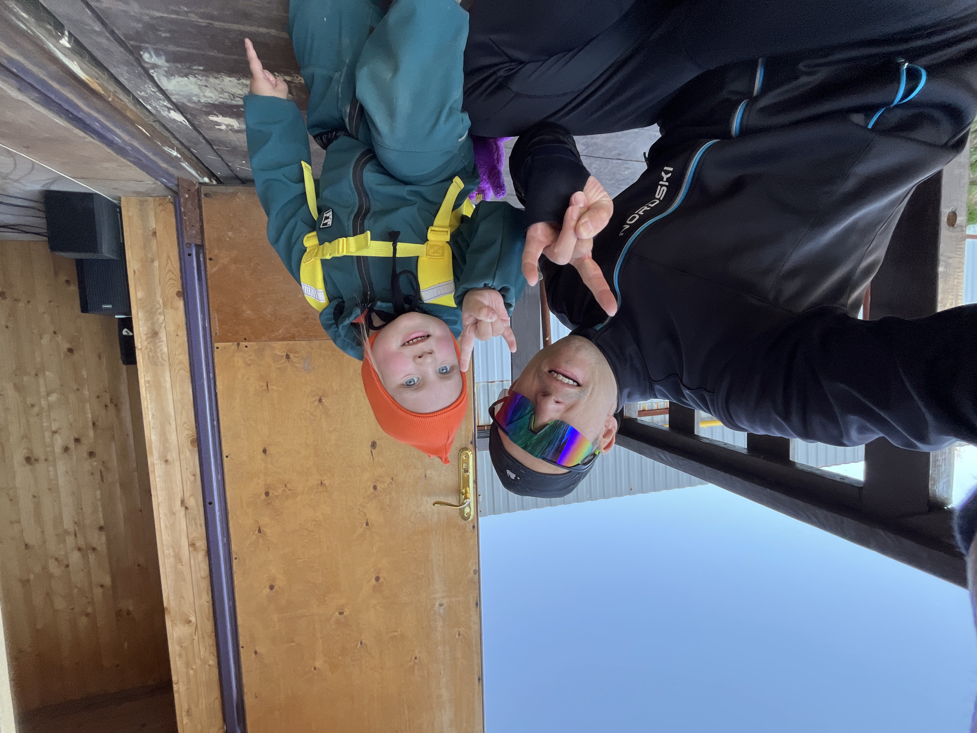 Two individuals are captured in a selfie; a man wearing multicolored sunglasses and a black cap, making a peace sign, and a young child with an orange cap and green snowsuit pointing upwards. The setting appears to be outdoors, with the sky visible in the background. There is a wooden structure or building nearby. The image doesn't provide enough information to identify the exact location in the city of Chaikovsky, Russia.