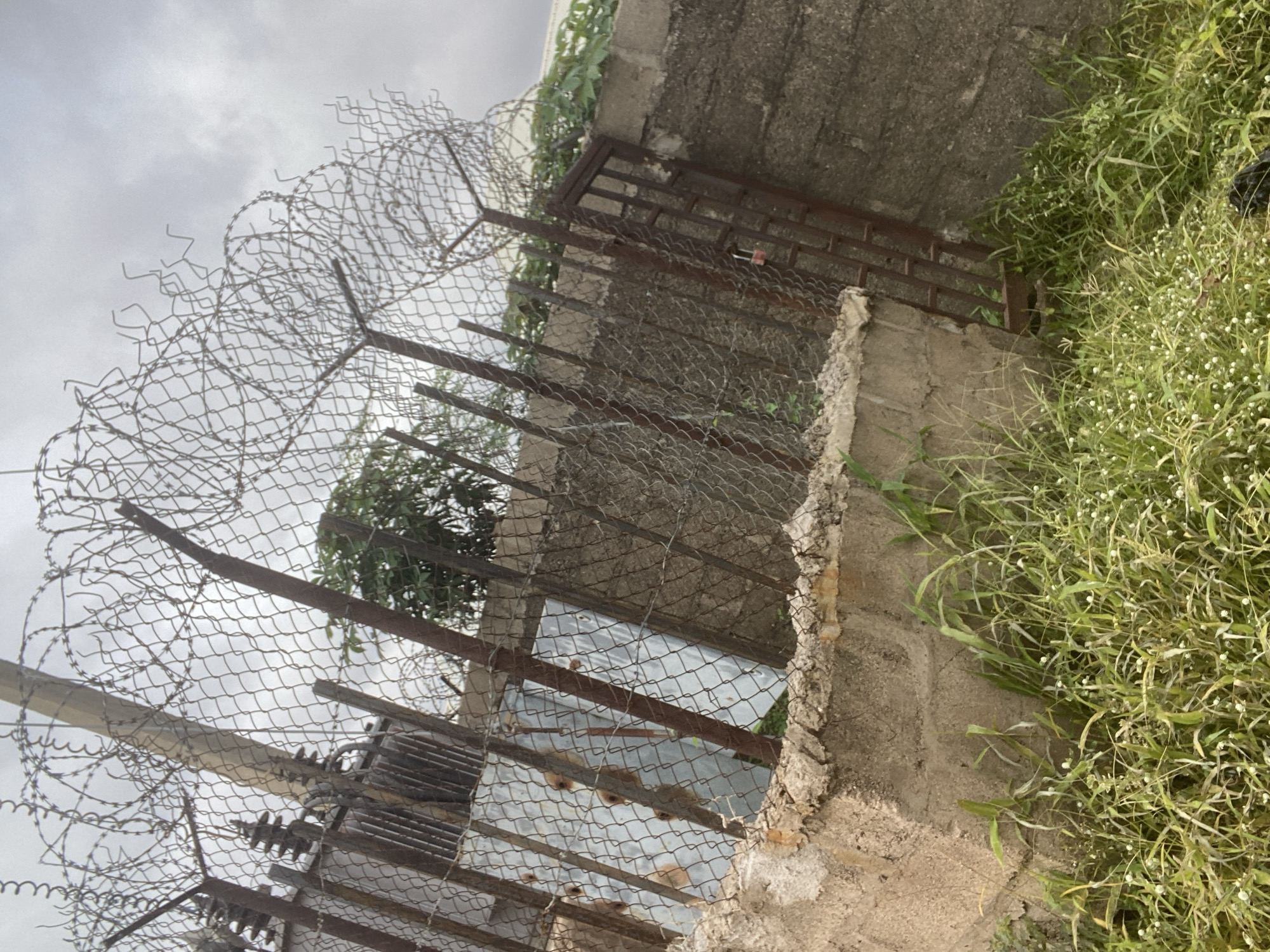 This image depicts a chain-link fence topped with barbed wire, constructed around a property. The fence is supported by concrete posts and rusty metal poles. Behind the fence, a building with a metal sheeting facade is visible, along with a rusty metal gate secured with a padlock. The area in front of the fence is overgrown with tall grass and some small white flowers. The scene appears to be in Gbaza, Nigeria, as indicated by the context. The sky is overcast with clouds, suggesting a possibly humid or rainy day. There are no people visible in the image.