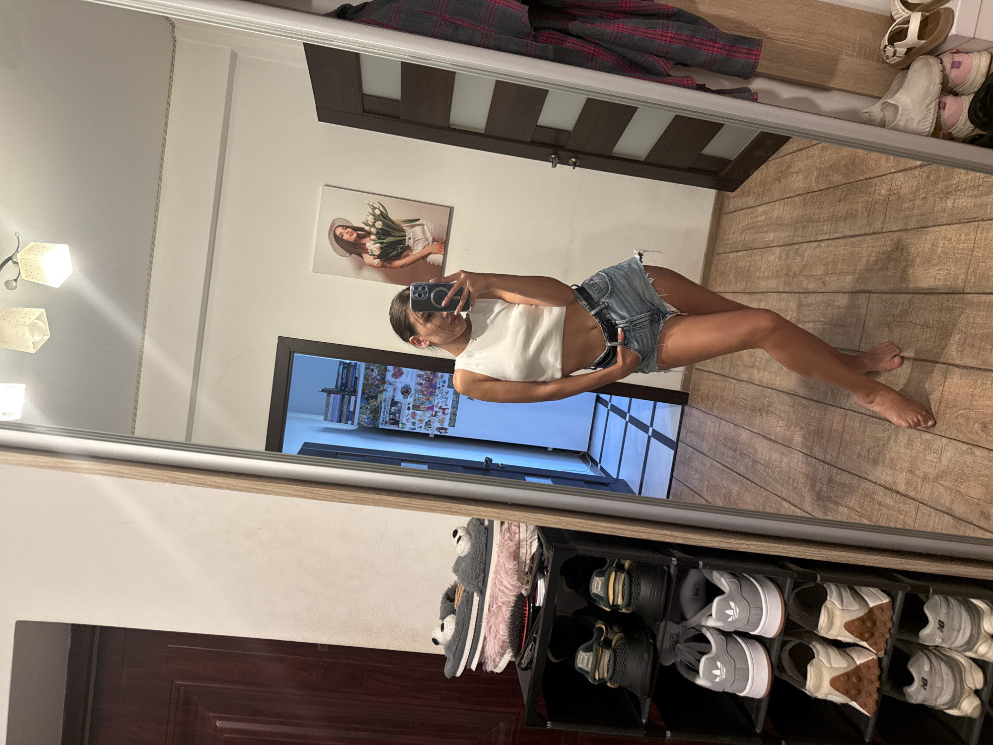 A young woman with dark hair is standing and taking a mirror selfie in what appears to be a hallway in Chișinău, Moldova. She is wearing a white crop top and denim shorts. Her phone is visible in her right hand. To her left, there is a shoe rack with several pairs of shoes and some slippers on top. In the background, there is a door and a framed picture on the wall. To her right, a door and a red and black plaid shirt are visible. The floor is made of wooden planks. The lighting in the room appears to be artificial.