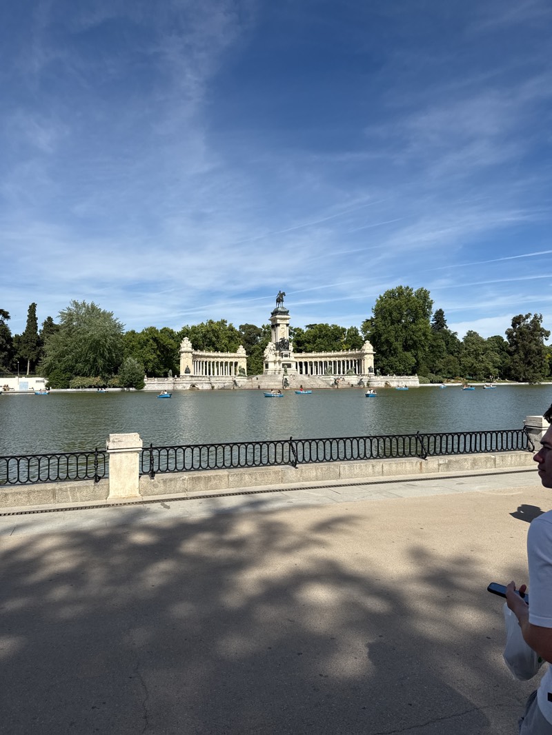 The image captures a vibrant, sunny scene in Madrid's El Retiro Park, specifically featuring the Monument to Alfonso XII overlooking the park's large lake. The central focus is the grand, white neoclassical monument with a semicircular colonnade, at the center of which stands a large equestrian statue of King Alfonso XII. Lush green trees frame the monument and line the distant shores of the lake.
The foreground shows a paved walkway with prominent dappled shadows from unseen trees, indicating bright sunlight. A sturdy black metal fence with stone posts separates the walkway from the calm waters of the lake. On the lake, several small rowboats with people can be seen enjoying the water, adding a sense of leisure and activity to the tranquil setting.
To the far right in the foreground, a person is partially visible, seen from the chest up, wearing a white t-shirt and holding a smartphone. The sky above is a clear, brilliant blue with a few wispy white clouds and several distinct contrails from aircraft, suggesting good weather. The overall impression is one of a pleasant, sunny day in a popular urban park. There is no visible text in the image.