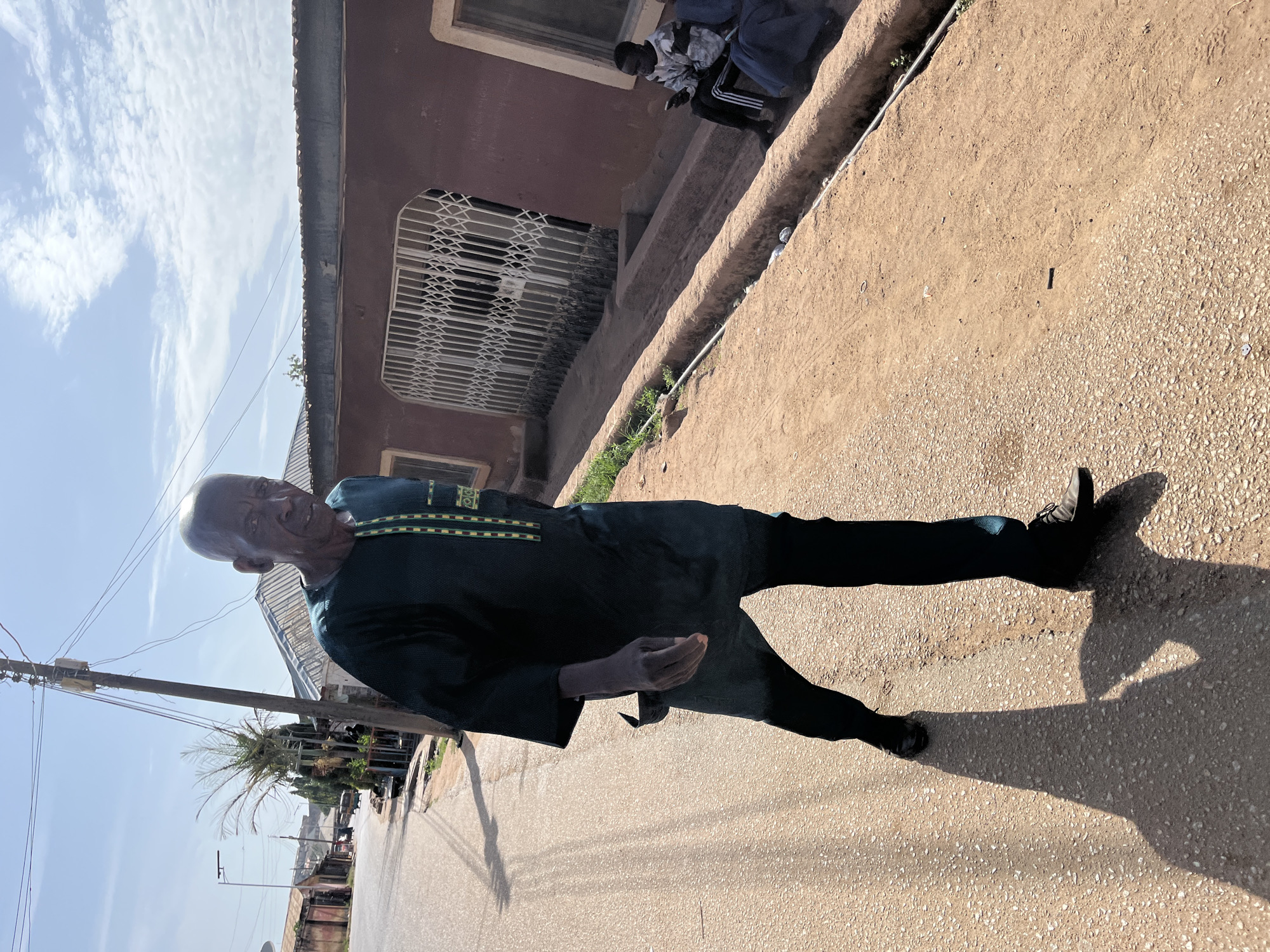 An elderly man is standing on the side of a paved road in Jos, Nigeria. He is wearing a dark green shirt with yellow and green trim around the collar and black trousers. The buildings visible are brownish, some with security bars on the windows. The weather appears sunny, based on the shadows cast. A person is sitting on a ledge or wall next to one of the buildings. Utility poles and wires are visible in the distance, suggesting a residential or commercial street.