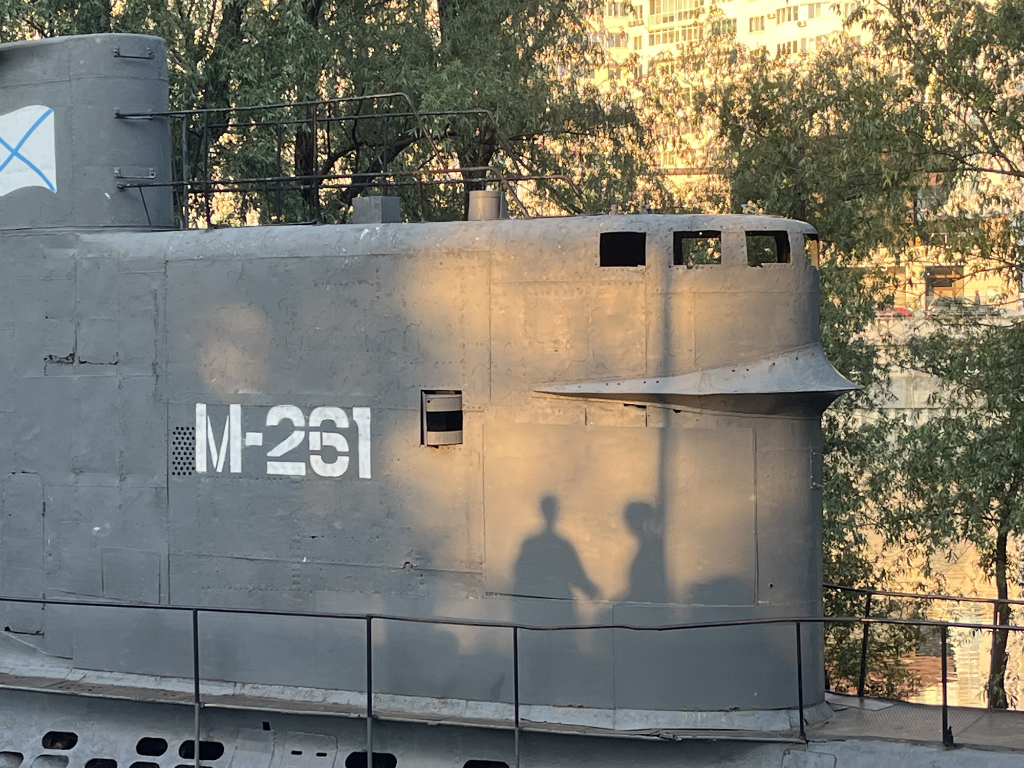 A side view of the conning tower of submarine M-261, a Soviet Project 613 (Whiskey-class) diesel-electric submarine, at its location in the city of Krasnodar, Russia, is seen. The submarine is painted gray. The number "M-261" is painted in white on the side of the conning tower. Two shadowed figures, possibly tourists, are projected onto the side of the conning tower. A metal railing runs along the length of the conning tower. Trees are visible in the background, with what appears to be a tall building beyond the trees.