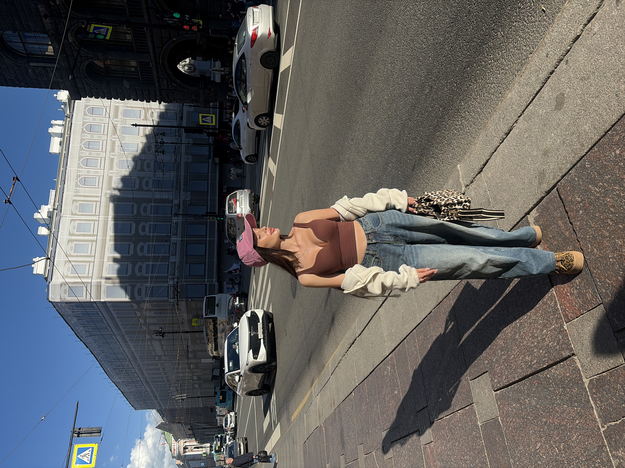 A young woman in a brown tank top, blue jeans, and a pink baseball cap is standing on a sidewalk in Saint Petersburg, Russia. She is wearing a cream-colored cardigan draped over her arms. The woman is holding a leopard-print purse. She is casting a long shadow on the granite sidewalk. Behind her, a street is lined with cars and buses, and a multi-story building with ornate architecture is visible. The sky is clear and blue, suggesting it is a sunny day. The time of day appears to be afternoon, given the long shadows. The overall mood seems relaxed and casual. Traffic lights and a pedestrian crossing sign are visible, indicating an urban environment. The building in the background may be undergoing renovation or is covered in scaffolding with a protective mesh.