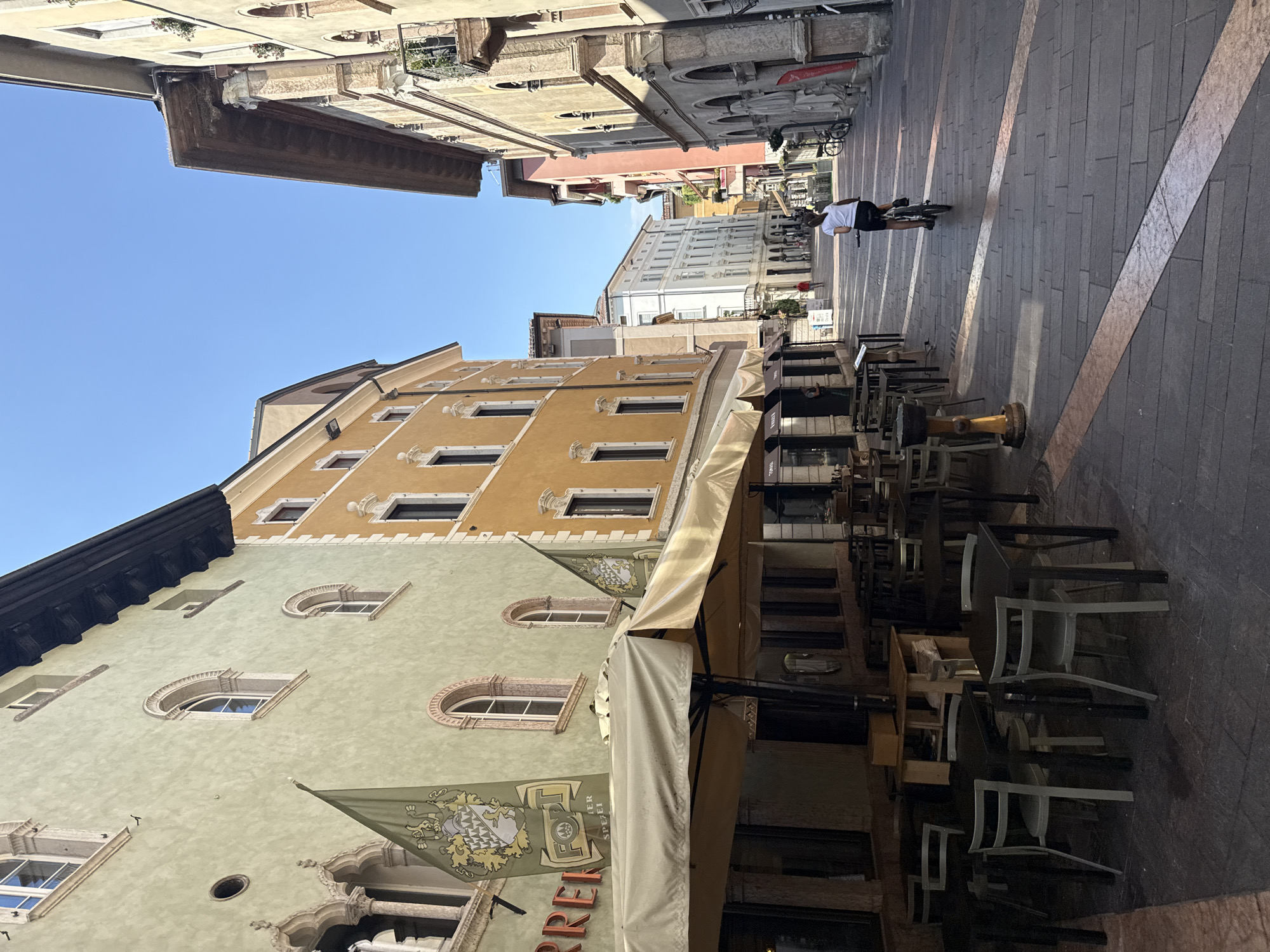 This image depicts a view down a narrow, European-style street in Trento, Italy, on a sunny day. The street is paved with stone, featuring decorative inlaid stripes. Buildings line both sides of the street, exhibiting distinct architectural styles. On the left, a building with a pale green facade features arched windows and decorative stone detailing. A large banner with a crest and text hangs from this building. In front of this building, an outdoor seating area for a cafe or restaurant is set up with numerous tables and chairs beneath a large, beige awning.
To the right of the street, buildings are painted in warmer tones, such as yellow and pink, with more elaborate window frames and balconies.
In the middle distance, a person is riding a bicycle away from the viewer down the street. Further down the street, other pedestrians can be seen walking. The sky is clear and blue. The overall impression is of a vibrant, historic urban setting during daytime.