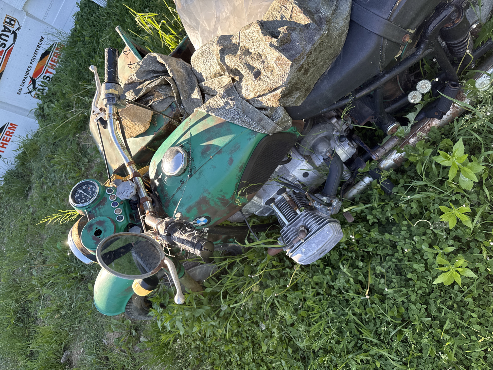 The image shows a partially disassembled green motorcycle lying on its side in a grassy area. A large rock and fabric scraps are resting on the motorcycle's fuel tank and seat. The motorcycle's engine is visible, as are gauges, handlebars, and a mirror. The immediate environment is characterized by green grass and weeds. In the background, there is a white box with the words "HAUSTHERM" printed on it. The location of the image is not discernible from the image's visual contents.