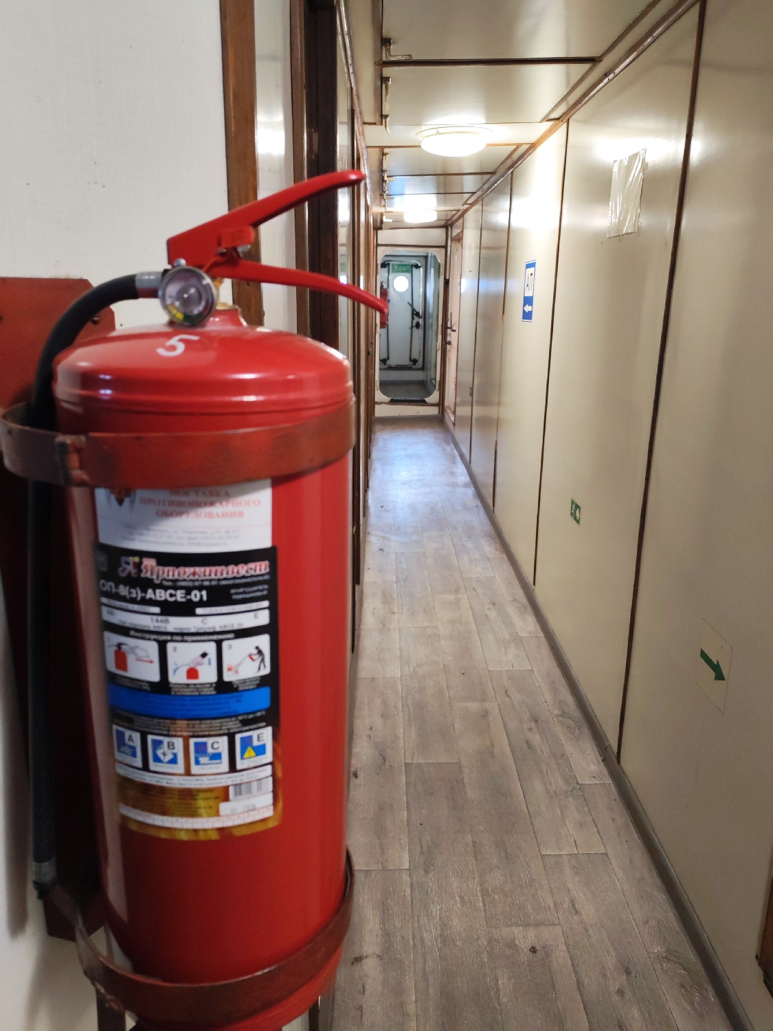 The image displays a functional, narrow corridor, likely on a ship or within an older building, viewed from a slightly elevated perspective. In the foreground, a large, bright red fire extinguisher is mounted on the left wall. It features a pressure gauge, a black hose, and a red handle, with the number "5" visible on its top. The extinguisher's label, partly visible, contains Cyrillic text reading "ПРОТИВОВОГО", "ОБОРУДОВАНИЯ", "*Приоритет", and "ОП-8(3)-ABCE-01". The label also includes instructional pictograms for use and symbols indicating fire classes A, B, C, and E.
The corridor extends into the background, illuminated by circular ceiling lights. The floor is covered with light gray, wood-patterned planks. The walls are paneled in a light cream or off-white color, separated by darker brown trim. Several doors line the right side of the corridor. Further down, an open doorway is visible, featuring a green "EXIT" sign above it. Other signs on the right wall include a blue square with a white arrow pointing right and a smaller green arrow near the floor also pointing right. No people are present, and the artificial lighting suggests an indoor setting where the time of day is not discernible.