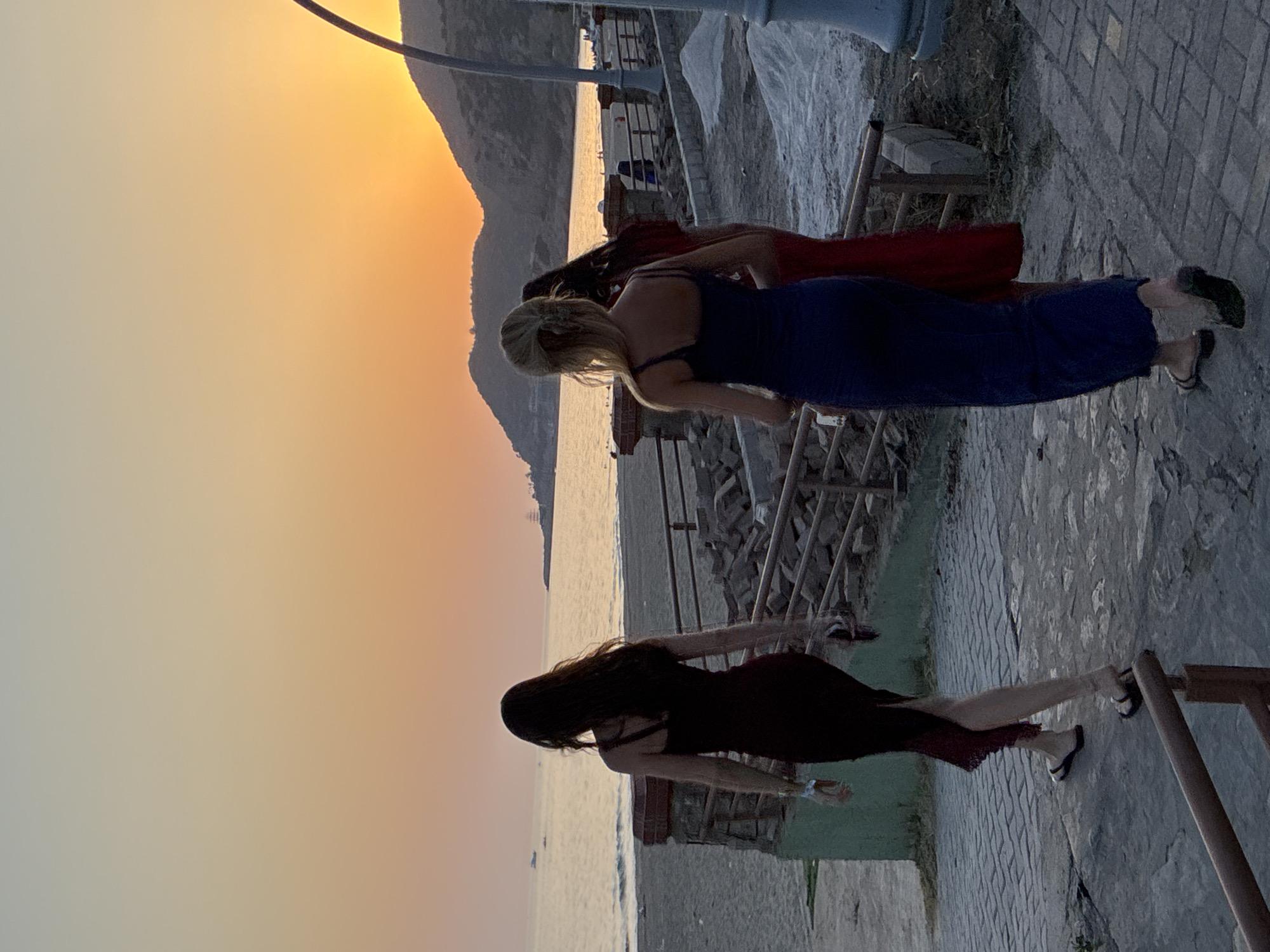 Three women are walking away from the camera on a paved walkway. They are dressed in long, flowing dresses. The woman in the foreground on the left is wearing a dark maroon dress. The woman in the middle is wearing a dark blue dress. The woman on the right is wearing a red dress. They are walking towards a beach and the sea. The sun is setting over the sea, casting a warm glow on the scene. In the distance, there are hills. The walkway appears to be part of a promenade in Alanya, Turkey, near the beach.