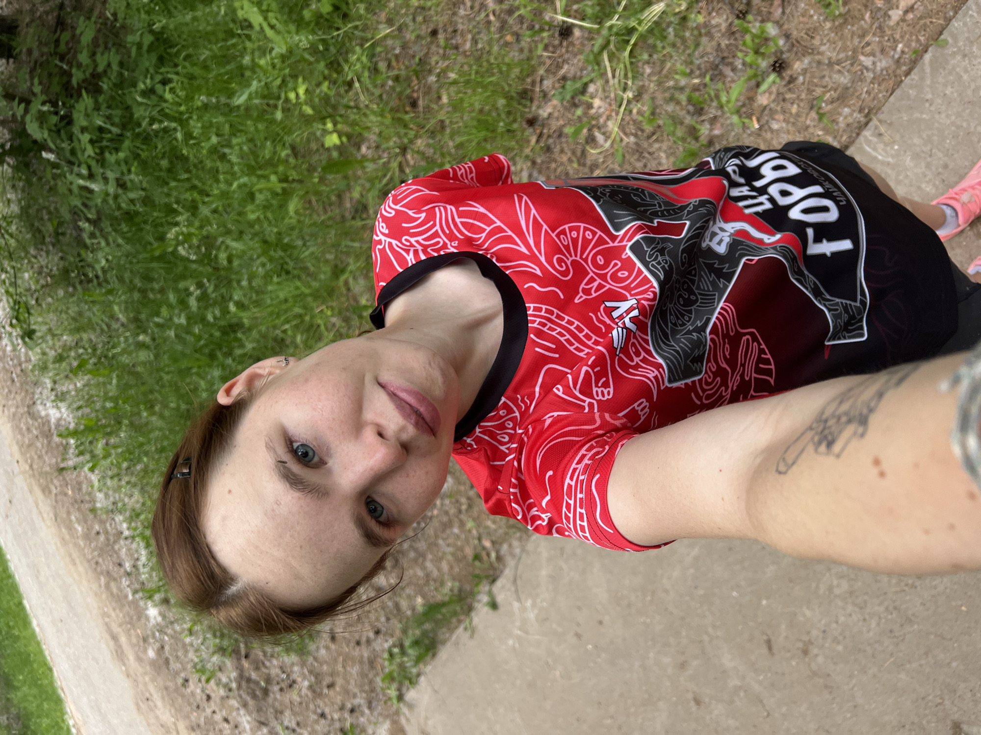 The image shows a young person taking a selfie outdoors, possibly while exercising. They are wearing a bright red and black athletic shirt with white graphic designs and some visible text. The person has light skin, blue eyes, and brown hair pulled back. Their expression is neutral, and they appear relaxed.
The background consists of greenery, a dirt path, and a paved surface, suggesting an outdoor setting such as a park or trail. The lighting indicates it is daytime, and the weather appears to be mild and possibly overcast due to the diffused light. The person's arm, with a visible tattoo, is in the foreground, along with their face and torso. They are also wearing pink athletic shoes.
The text on the shirt appears to be in Cyrillic. Partially visible text on the black section of the shirt includes "TOPB". The red portion of the shirt features intricate white line designs.
The image was taken in Chaikovsky, Russia, implying the person may be participating in outdoor activities specific to that region. The overall impression is one of casual outdoor activity.