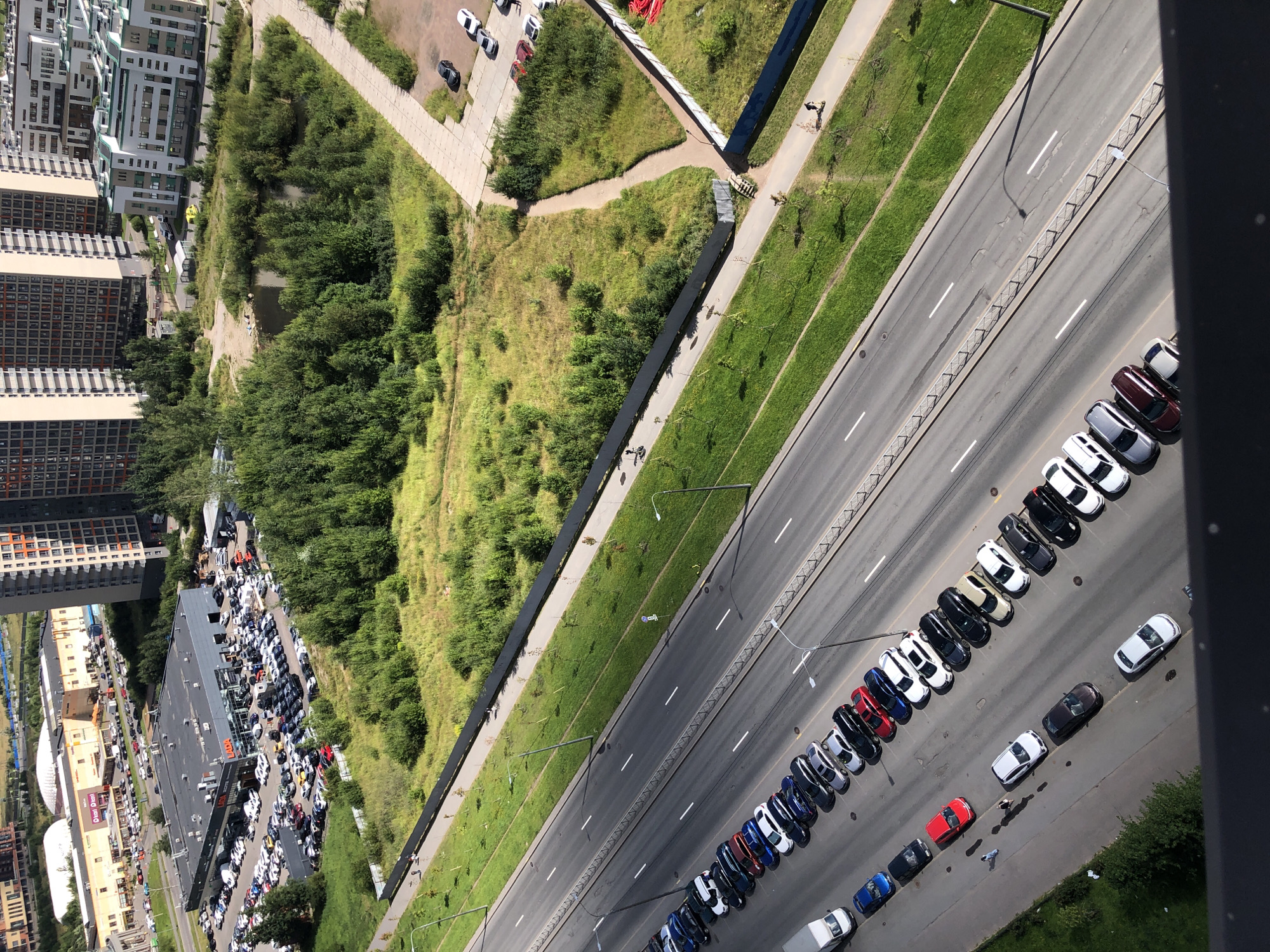The aerial photograph shows a section of a multi-lane road in a city, with a parking lot filled with vehicles alongside it. To the left of the road, a large car dealership, identified by a "LADA" sign, is visible, with many cars parked in its lot. Across the road, a green, overgrown field with a small pond and scattered trees occupies a significant portion of the frame. In the background, several modern, multi-story buildings, some with distinctive architectural features, dominate the skyline. A paved walkway runs along the edge of the field, with a few pedestrians visible. The road itself is lined with parked cars.