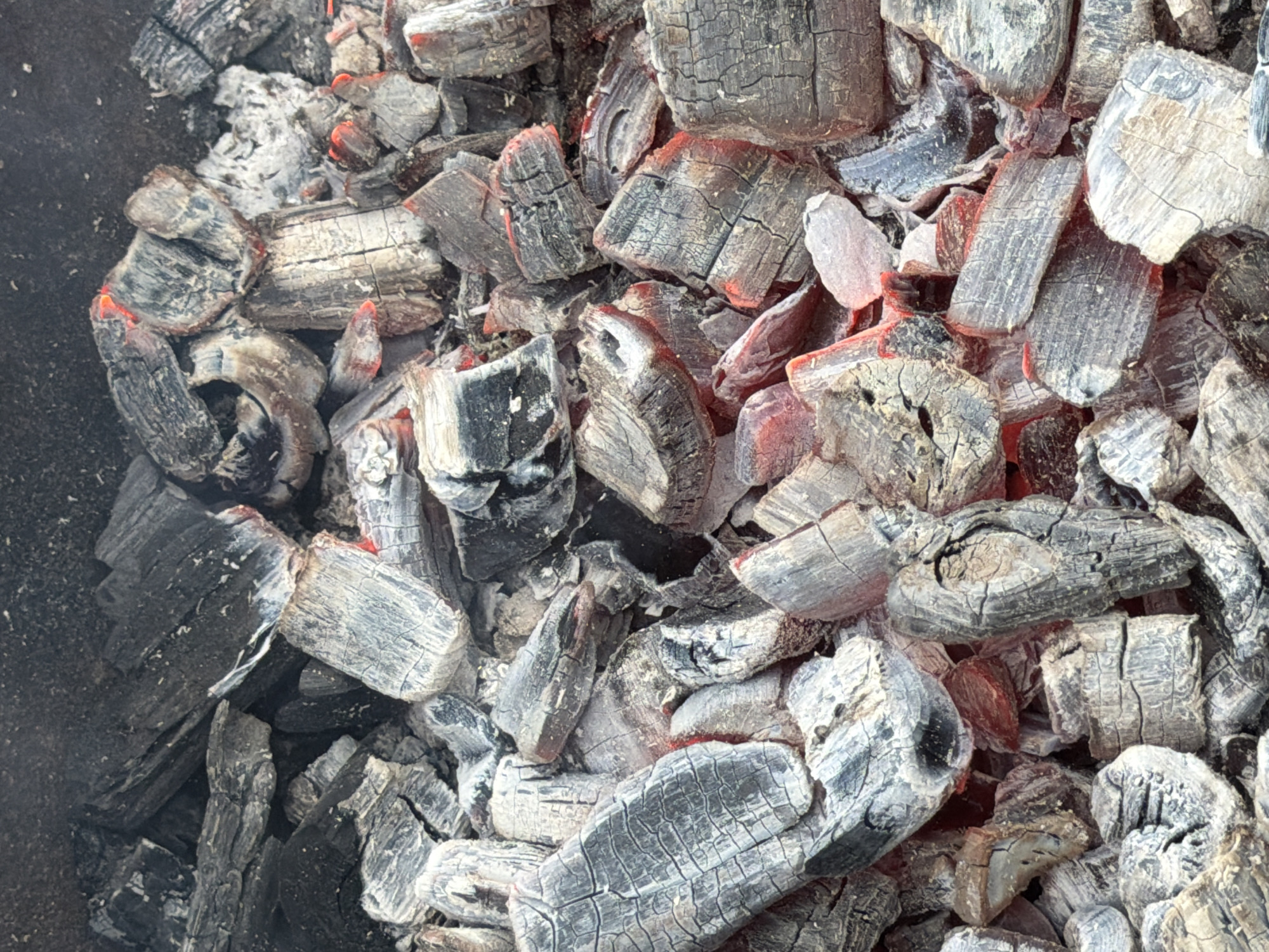 The image shows a pile of partially burnt charcoal briquettes. The briquettes are primarily black and gray with some red embers glowing. The charcoal has a blocky texture with cracks and fissures. Some areas show white ash accumulation. The scene appears to be contained within a dark-colored container, potentially a grill or fire pit. There is no indication of people or actions in the image. The location is not identifiable from the image.