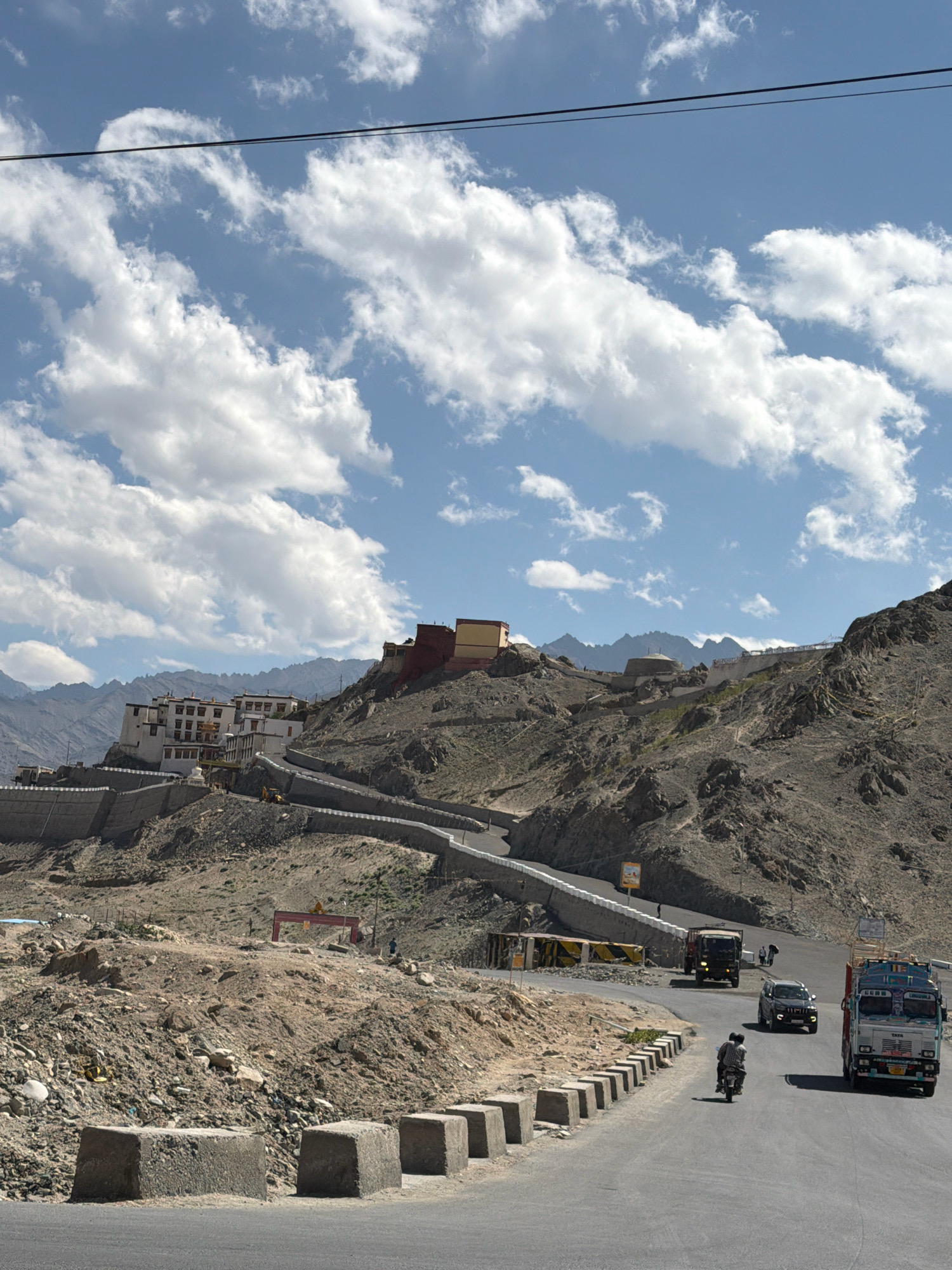 The image captures a scenic view of the Spituk Monastery, located near Leh in Ladakh, India. The monastery complex, characterized by its white and red-colored buildings, is perched on a hilltop overlooking a winding road. Several vehicles, including trucks, a car, and a motorcycle, are seen navigating the road. The surrounding landscape is rugged and mountainous, with sparse vegetation. The sky is a clear blue, dotted with fluffy white clouds. The presence of a power line across the top of the frame suggests the area is connected to electricity. The scene depicts a typical day in this high-altitude region, with transportation and architectural heritage coexisting in a dramatic natural setting.
