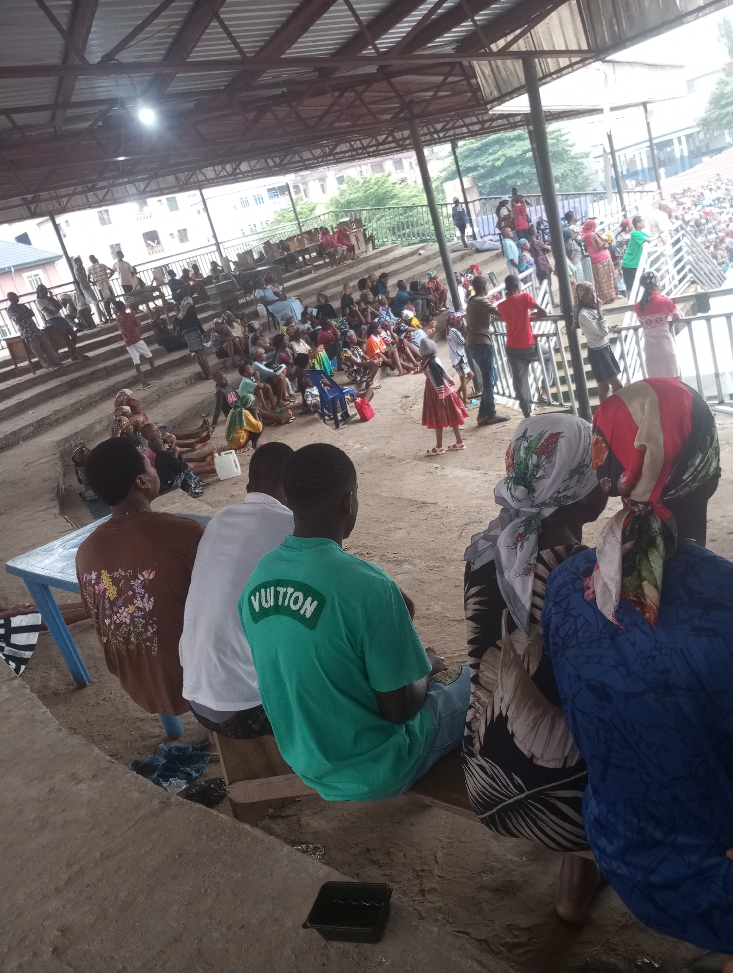 The image shows an outdoor gathering of people under a covered structure with a corrugated metal roof supported by metal beams and columns. The structure appears to be an open-air pavilion or bleacher area. Many people are seated or standing on tiered steps that rise from the ground level. On the ground level, several individuals are seated. A person in a green t-shirt with the word "YUITTON" printed on the back is prominently in the foreground, facing away from the camera. To their left, another person in a white t-shirt is also facing away. Further to the left, a person in a brown t-shirt with a floral pattern is seated. A blue plastic table is situated near the front of the seating area. The background reveals buildings and trees. The ground is dusty or dirt-covered. The overall atmosphere suggests a community event or gathering.