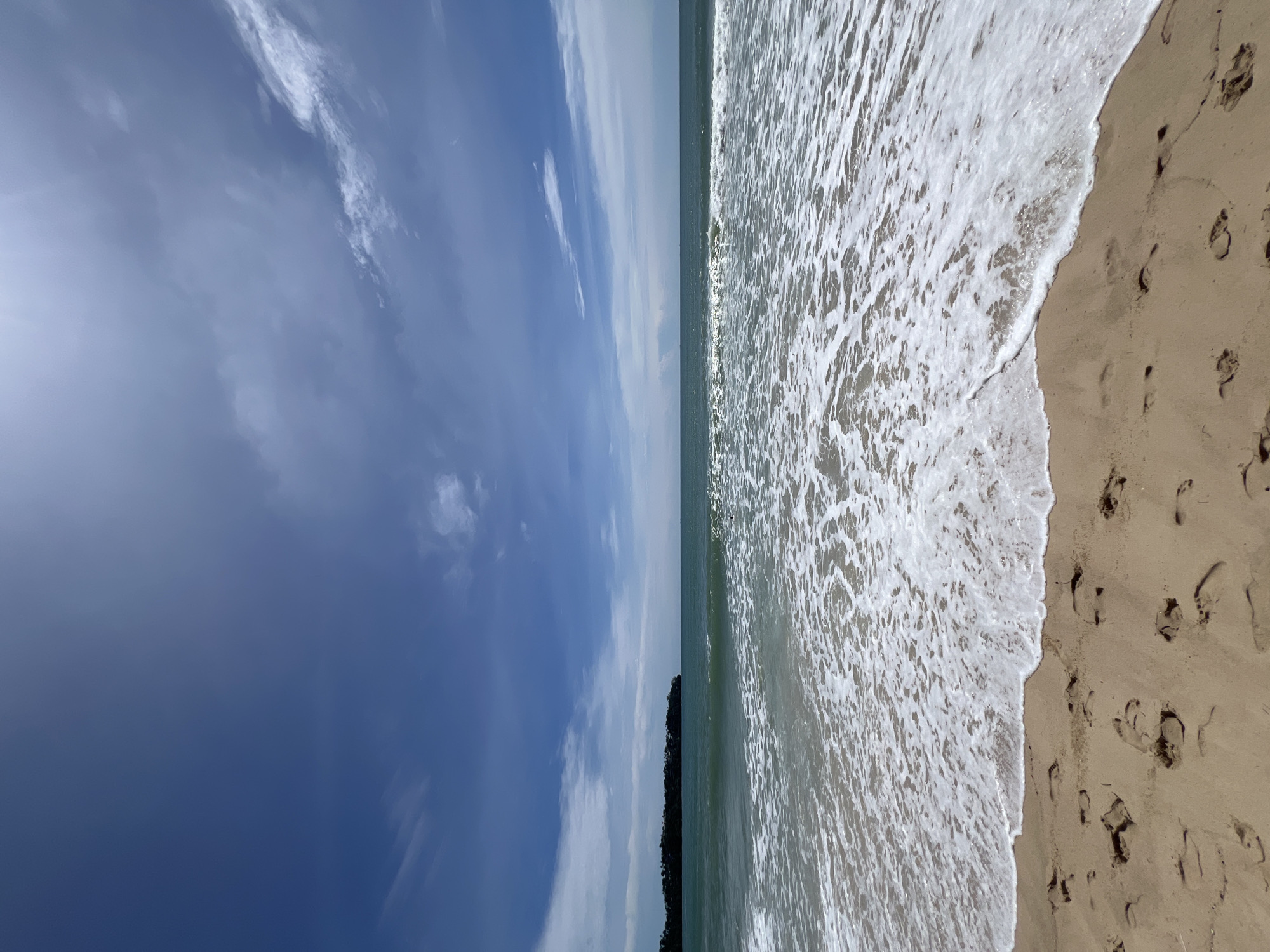 The image presents an outdoor scene at a beach in Unknown, Unknown, under a bright, sunny sky. The upper approximately two-fifths of the frame consists of a predominantly clear, vivid blue sky with scattered white, wispy clouds, particularly concentrated in the mid-right and mid-left areas. A bright light source, presumably the sun, is positioned high in the sky, causing a glare in the upper right corner. The horizon line is flat and well-defined, separating the sky from the ocean.
The middle section of the image, extending from the horizon to the breaking waves, features the tranquil ocean with a distinct teal or greenish-blue hue. On the far left, a dark green, low-lying landmass or island covered in dense vegetation is visible, extending partially into the sea. Several lines of gentle waves are present, with the most prominent one breaking into a continuous expanse of white, frothy foam across the lower-middle section of the image.
The bottom approximately one-third of the frame displays a sandy beach. The sand is a light tan color and is conspicuously marked with numerous human footprints of varying sizes and orientations, indicating recent pedestrian activity. The sand directly at the water's edge appears darker due to wetness. No individuals or man-made objects other than the footprints are visible.