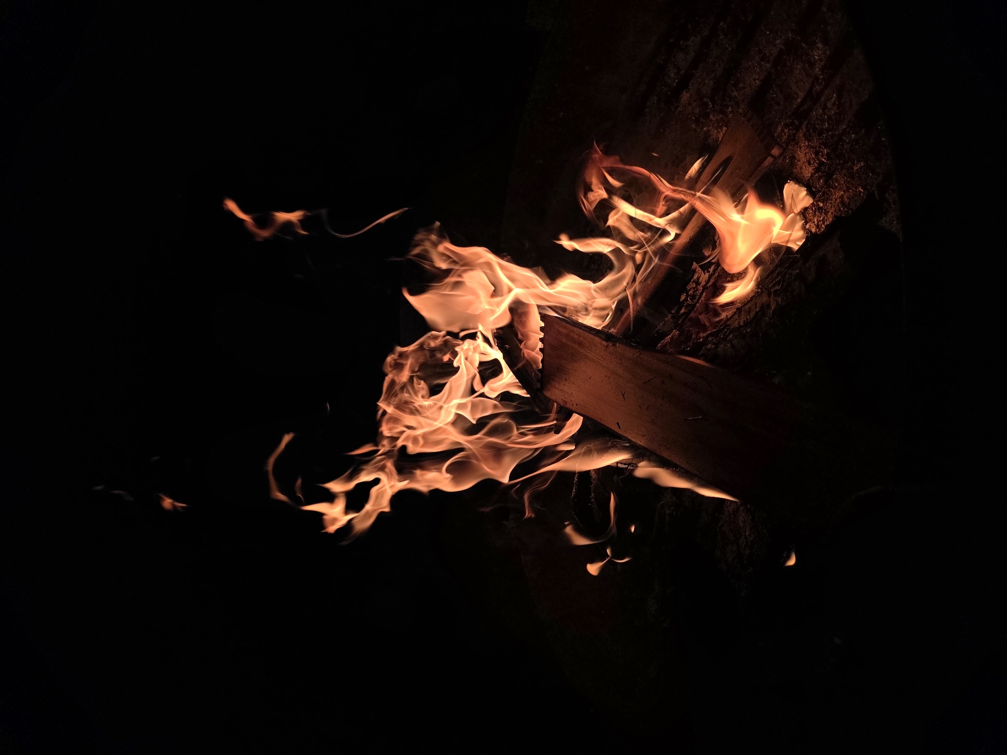 A close-up, low-angle, vertical shot captures a bonfire in the dark, with flames licking upwards from burning logs. The fire is a vibrant mix of orange and yellow hues, with wisps of smoke curling into the blackness of the night sky. The logs themselves appear charred and are partly obscured by the intense heat and light of the flames. The scene is devoid of any people. The location of this bonfire is not specified within the image itself, therefore it is impossible to determine if it is in Строитель-нефтяник Снт, Russia.