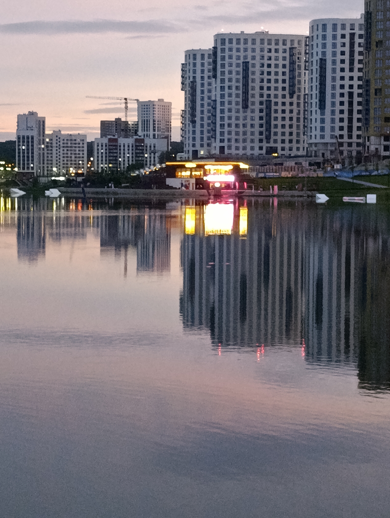 The image depicts a waterfront scene at dusk in Penza, Russia, likely in the area of the "Art Space" complex on the embankment of the Sura River. A series of modern, multi-story residential buildings with numerous windows are prominent on the horizon. In the foreground, a body of water reflects the buildings and the softly lit sky. A building with brightly illuminated windows, possibly a cafe or restaurant, is situated along the waterfront, its vibrant lights casting a strong reflection on the water's surface. The overall atmosphere is calm and serene, characteristic of an evening in a city.