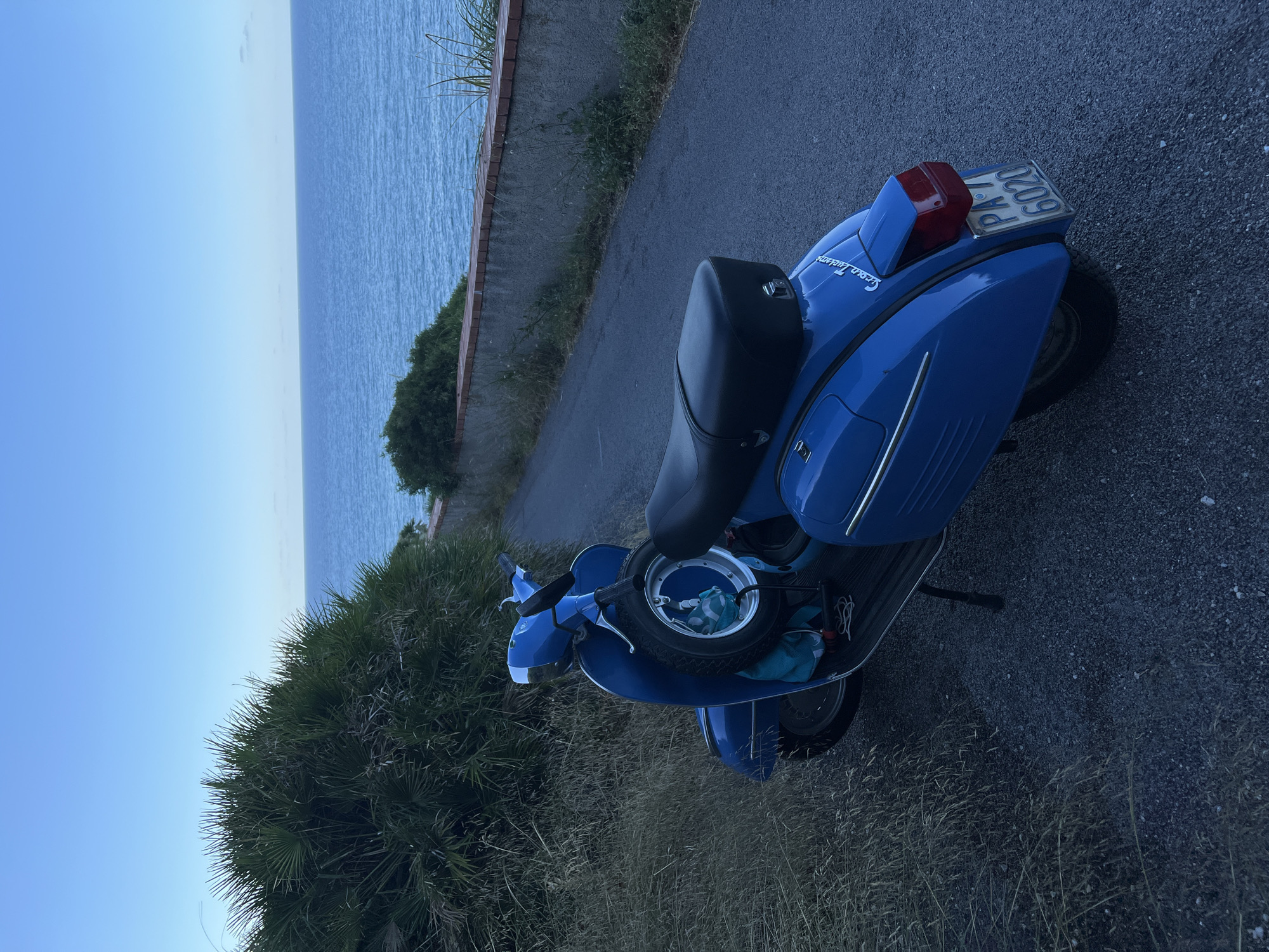 A blue vintage Vespa scooter is parked on a paved road next to a grassy embankment. The scooter is positioned at an angle, facing away from the viewer and slightly to the left. It has a spare tire mounted on the front. In the background, a body of water, likely the sea, stretches to the horizon under a clear blue sky. A low, weathered wall runs along the edge of the road separating it from the embankment and the sea. The scene appears to be in the town of Trabia, Italy, a coastal municipality in the Metropolitan City of Palermo, Sicily. The overall mood is serene and peaceful, suggesting a quiet moment at dusk or dawn.