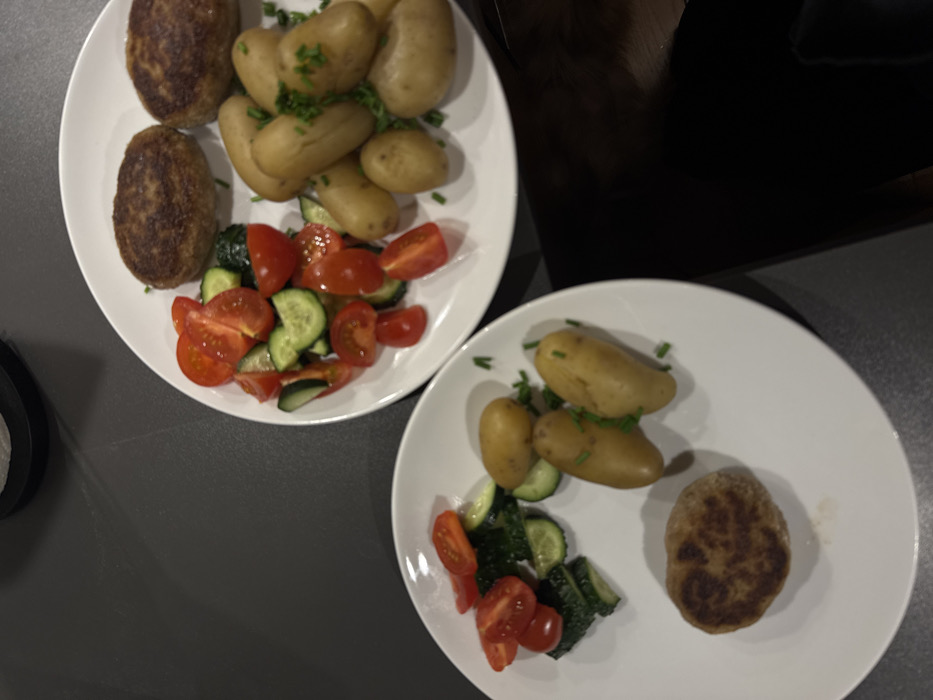 Two white plates rest on a dark gray surface. Each plate contains a meal. The meal consists of boiled potatoes, slices of cucumber and tomato, and a patty resembling a meatloaf. The plate on the left features more patties and a larger portion of potatoes. Fresh herbs are sprinkled over the potatoes. The food appears freshly prepared.