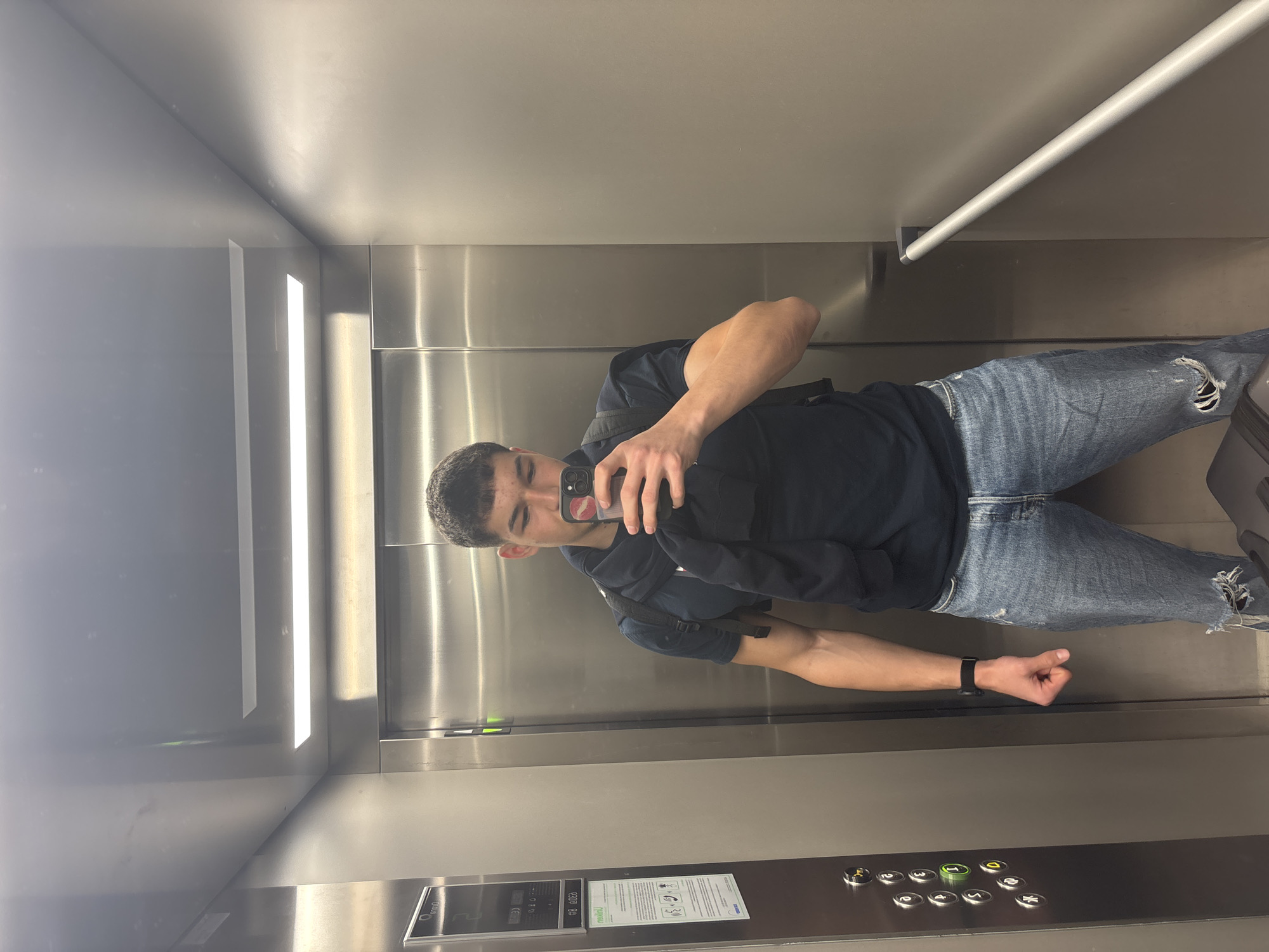 A young man is taking a selfie in what appears to be an elevator. The interior of the elevator is stainless steel, with a horizontal grab bar visible at the top. The man is wearing a dark-colored hoodie, blue jeans, and a watch on his left wrist. He is holding a smartphone in front of his face, taking the photo. There is a panel of elevator buttons near the bottom of the frame. The location cannot be determined to be Plovdiv, Bulgaria based on the image alone.