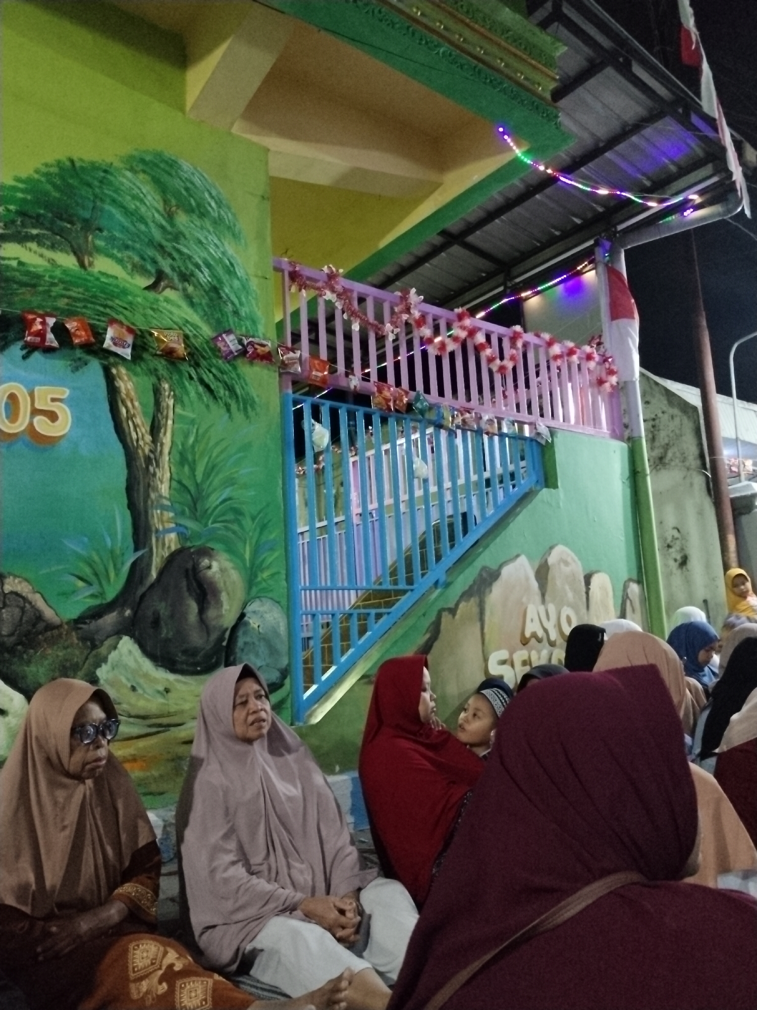 The image depicts an outdoor gathering of people in what appears to be a community space in Ditotrunan, Indonesia. In the foreground, several individuals are seated on the ground, some wearing hijabs and traditional Indonesian attire. To the left, a mural on a green wall features a stylized tree and rocks, with the number "05" prominently displayed. Behind the seated individuals, a colorful painted staircase with blue and purple railings leads up to a balcony adorned with festive red and white decorations and string lights. The background shows a corrugated metal roof and a wall, suggesting a simple building structure. A partial Indonesian flag is visible on the right side of the frame. The overall scene suggests a local event or celebration taking place in the evening, indicated by the lighting and the presence of decorative lights.