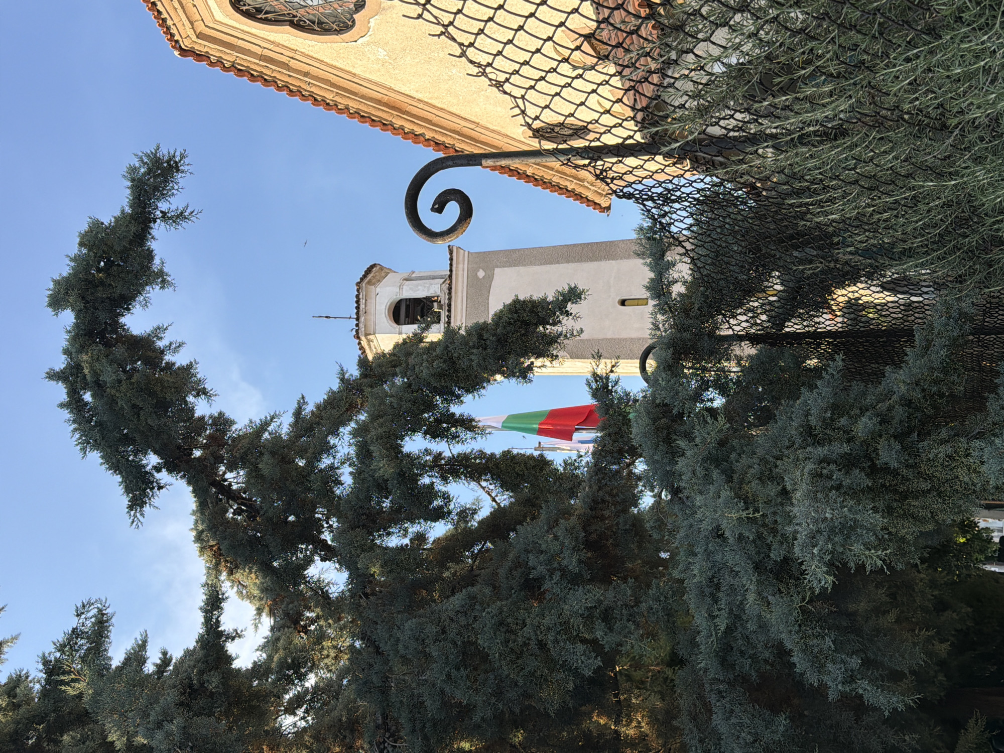 The image captures a Bulgarian flag flying in front of a church tower in Pazardzhik, Bulgaria. The flag, displaying its national colors of white, green, and red, is partially obscured by the branches of a large evergreen tree in the foreground. Behind the flag, a tall, light-colored church tower with a bell is visible against a clear blue sky. To the right of the tower, part of the church building is seen, with an arched window and a terracotta tiled roof. A black metal fence runs across the lower part of the image, with a decorative spiral element on a post in the mid-right. Rosemary bushes are growing along the fence. The scene appears to be set on a sunny day, with the sunlight casting shadows and highlighting the architectural details. There are no people visible in the image.