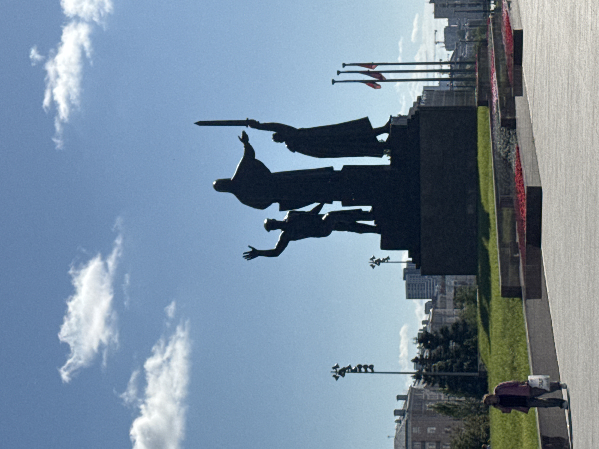 The image shows a large bronze statue of three figures, two standing and one in the center slightly elevated, holding a sword aloft. The statue is silhouetted against a bright blue sky with scattered white clouds. In the foreground, a paved area leads to a grassy slope with a flowerbed. To the left, a lone person walks, carrying a white bag. In the background, buildings and trees are visible, along with lampposts and flags. This is the "Perm City Defenders" monument, located on Komsomolsky Prospekt in the city of Perm, Russia.