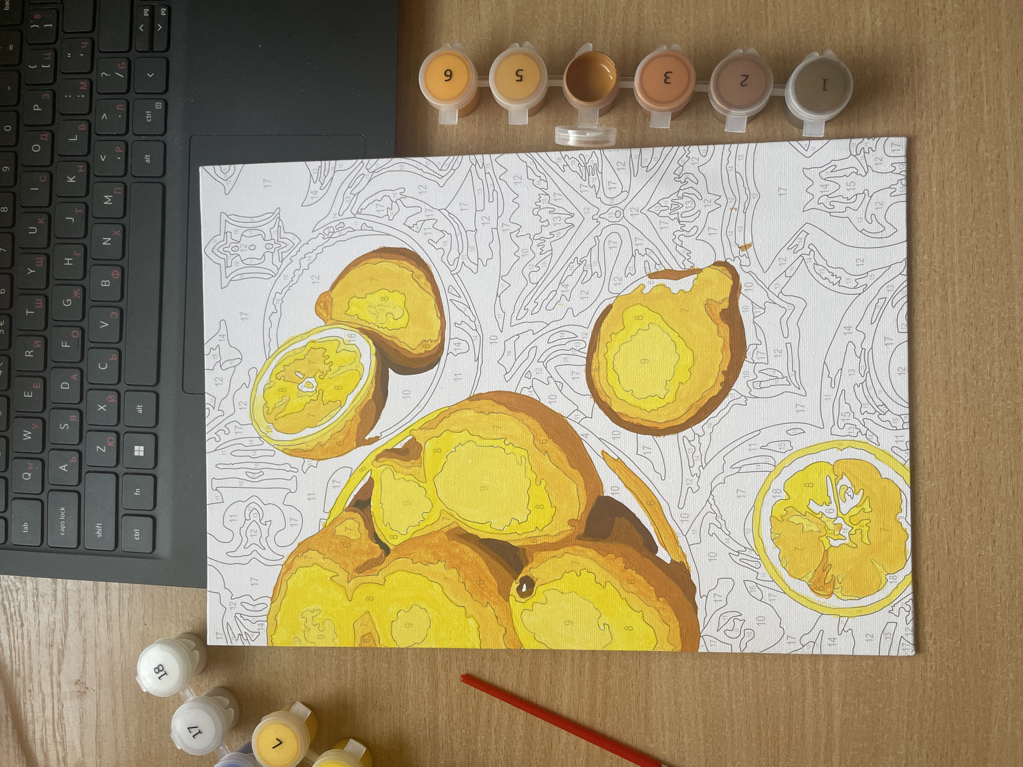 An overhead view captures a creative workspace featuring a partially completed paint-by-number artwork, a laptop, and painting supplies, set against a wooden surface.
The central subject is a paint-by-number canvas depicting several lemons or similar citrus fruits, some whole and some sliced. Many sections of the fruits are already painted in shades of yellow and orange, corresponding to numbers like "6," "8," "9," and "10" visible on the canvas. The background and unpainted areas are white, showing outlines and numbers awaiting color.
Arranged at the top and bottom of the canvas are small plastic paint pots with numbered lids, indicating different colors. Visible numbers on the lids include "1," "2," "3," "5," "6," "9," "17," and "18." A red paintbrush rests at the bottom, near other paint pots.
To the left, a black laptop keyboard is partially visible. It features both Latin (QWERTY layout) and Cyrillic characters, such as "Ы," "Д," "Ж," "Э," "Ч," "Ю," and "Я," common in countries like Bulgaria. Keys like "tab," "caps lock," "shift," "ctrl," "fn," "alt," "PgUp," "PgDn," and "backspace" are discernible.
The scene suggests an individual engaged in a leisure activity or hobby, specifically painting, in an indoor setting. The presence of the Cyrillic keyboard is consistent with the provided location context of Plovdiv, Bulgaria.