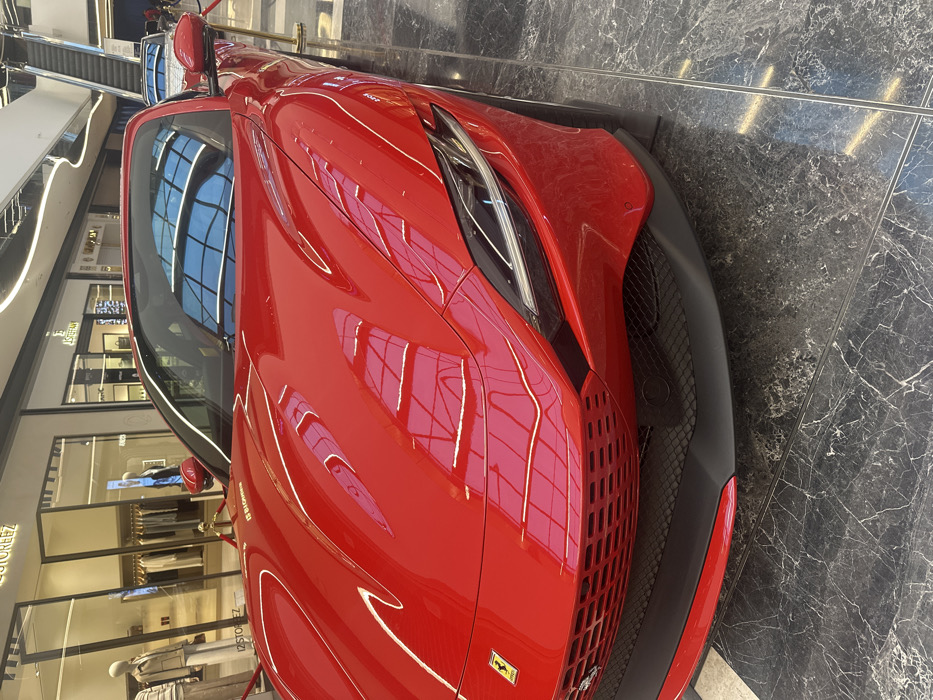 A red Ferrari Roma sports car is prominently displayed on a dark, polished, veined floor within a modern, multi-level indoor shopping mall in Tashkent, Uzbekistan. The vehicle's front section, including the hood, distinct grille, and right LED headlight, dominates the foreground. The highly reflective red bodywork exhibits clear reflections of the mall's ceiling architecture, overhead lighting, and a prominent blue, grid-patterned ceiling or skylight structure on the windshield and upper body. To the left, several glass-fronted retail stores are visible, one labeled 'ISTOREEZ' and another 'J.STEFAN', featuring clothing on mannequins. Gold-colored stanchions with red ropes partially enclose the display area around the Ferrari. An escalator ascends in the background on the right, and a portion of a second, lighter-colored vehicle is visible behind the displayed Ferrari. No individuals are clearly depicted in the immediate vicinity or interacting with the vehicle.