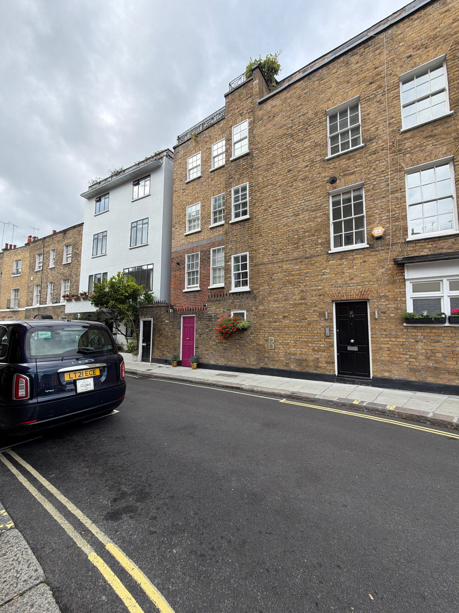 The image depicts a street scene in London, United Kingdom, under an overcast sky. In the foreground, a dark blue London taxi is parked on the left side of the asphalt road, which is marked with double yellow lines. The taxi has a visible license plate with the characters "LT21 ECE". The background features a row of terraced buildings with brick facades of varying colors. The building directly behind the taxi is painted white and has multiple windows. To its right, a taller building constructed of yellow brick has several windows and a black door. Further to the right, another brick building has a vibrant pink door. Planters with red flowers are visible on some window ledges. The environment appears to be a residential street.