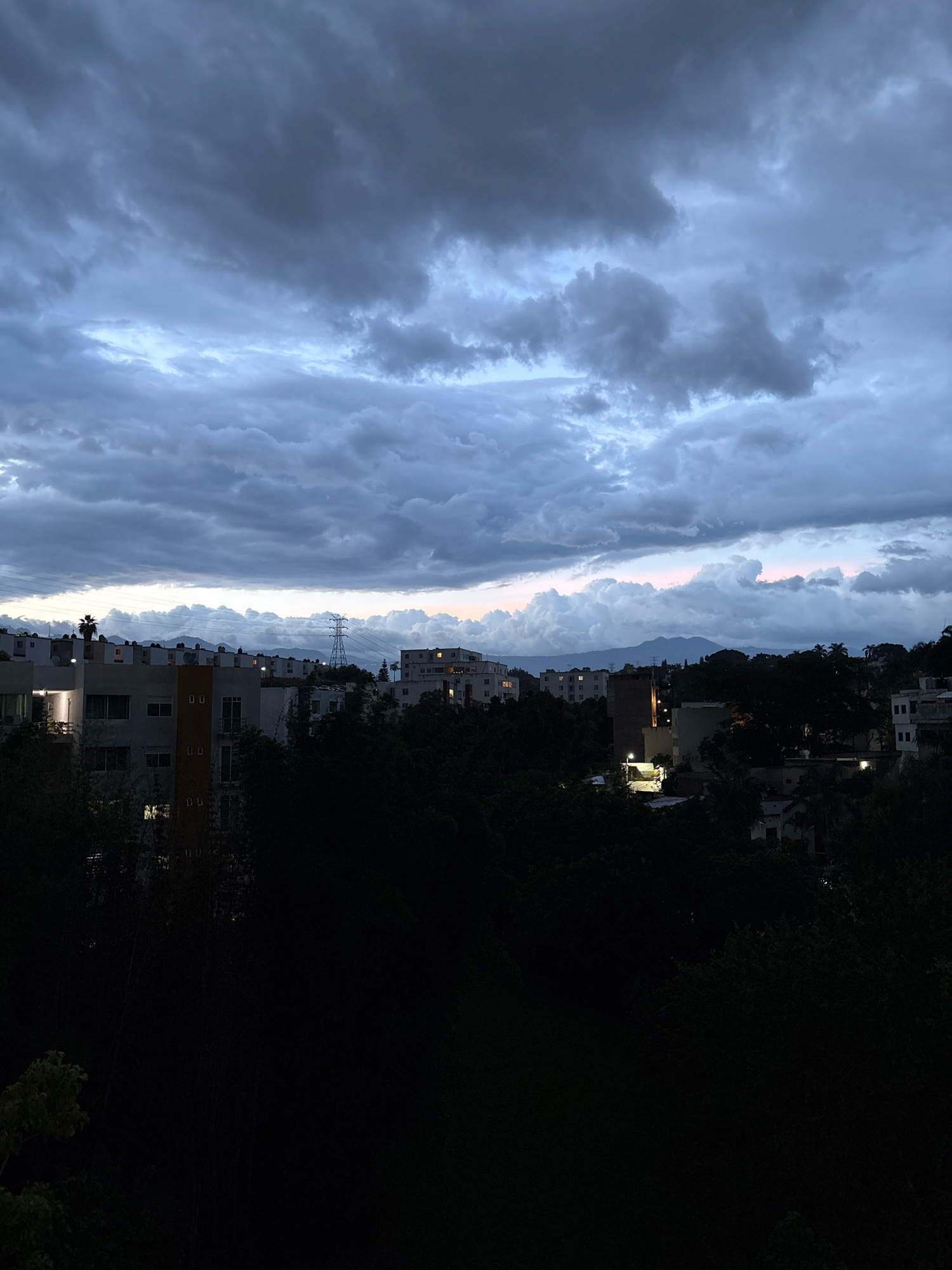 The image depicts a city skyline at dusk, with a dramatic, cloudy sky overhead. The clouds are a mix of dark grays and lighter blues, with hints of pink and orange near the horizon, suggesting the last rays of sunlight. Below the sky, a dense layer of dark green trees partially obscures the view of buildings.
Several multi-story buildings are visible, some with illuminated windows, indicating activity within. The architecture appears to be modern residential or commercial structures. In the mid-ground, a tall electrical tower stands out against the sky, and power lines stretch across the landscape. In the distance, mountains form a hazy backdrop.
The foreground is dominated by dark, silhouetted foliage, making it difficult to discern specific details. The overall impression is one of an urban area transitioning into night, with a somewhat somber or atmospheric mood due to the heavy cloud cover and low light conditions. The image was taken from an elevated vantage point, looking out over the city. The scene is consistent with a residential or mixed-use area in Cuernavaca, Mexico, given the mountainous topography and typical architectural styles observed in such cities.