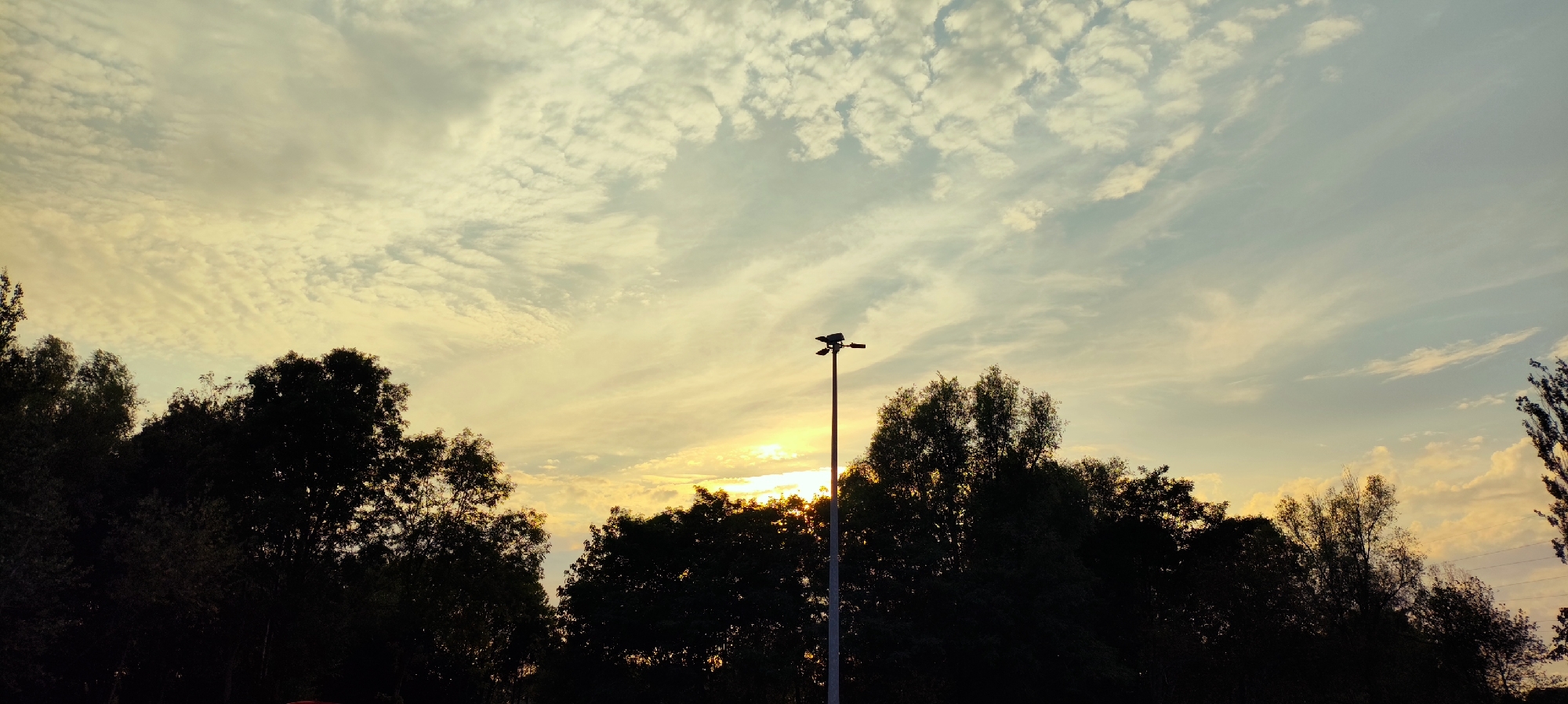The image depicts a sunset scene with a dark silhouette of trees lining the bottom half of the frame. A tall, grey lamppost with two lights is positioned slightly off-center to the right, cutting through the tree line. The sky is filled with wispy, golden-yellow and pale blue clouds, indicating the sun is low on the horizon and setting. The overall impression is of a tranquil evening in an outdoor setting, possibly a park or a suburban area within Knowsley, United Kingdom, given the lamppost and tree-lined landscape. There are no people or discernible actions visible in the image.
