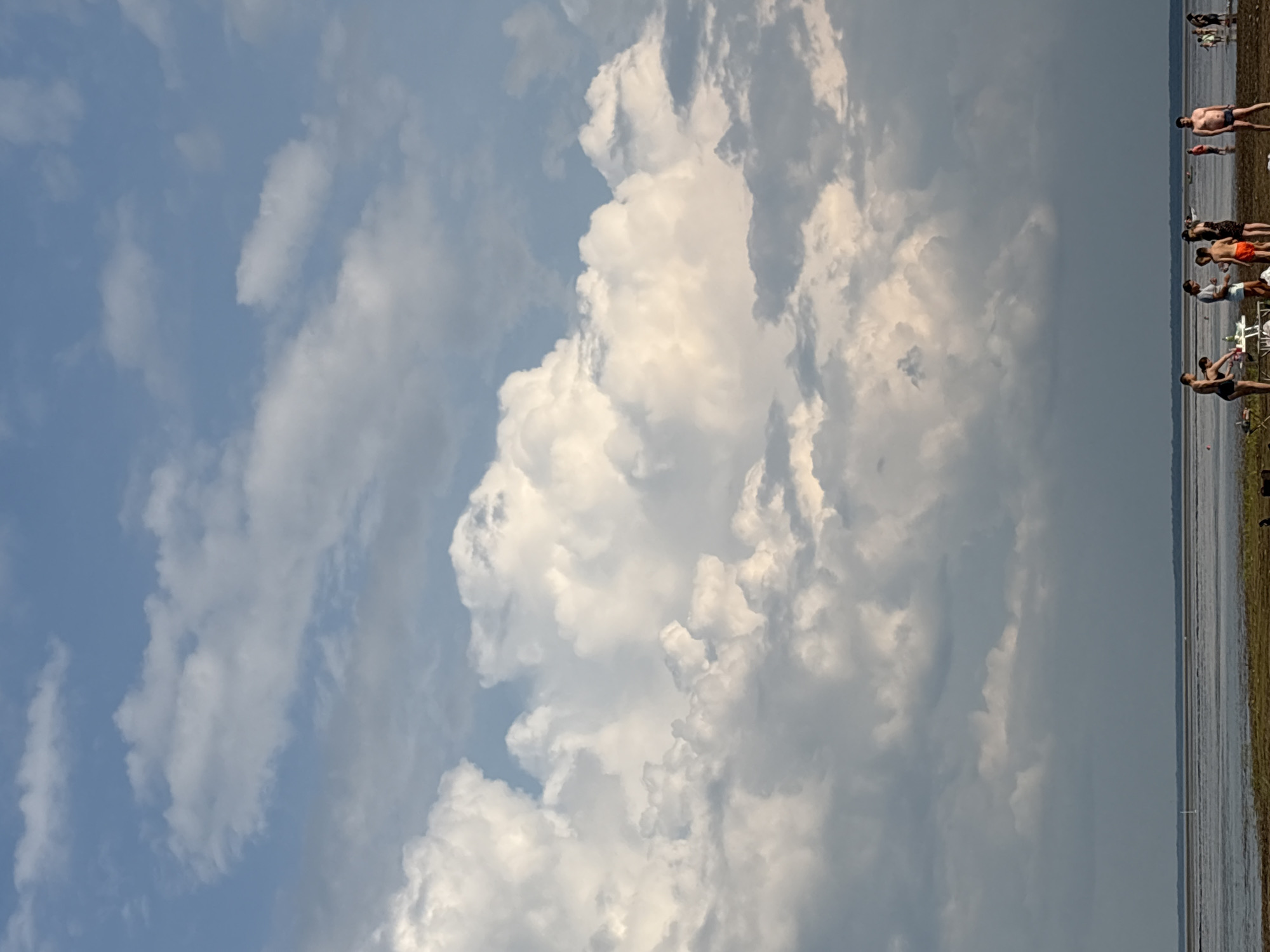 The image depicts a group of people on a beach in Koshkino, Russia. The sky is filled with large, dramatic clouds, some of which are illuminated by sunlight, while others are darker and appear to be gathering. A calm body of water stretches across the lower portion of the image, with a thin strip of land and beach in the foreground. Several individuals are present on the beach, some standing and interacting, while others appear to be engaged in activities near a picnic table. The overall scene suggests a leisurely day by the water, under a dynamic sky.