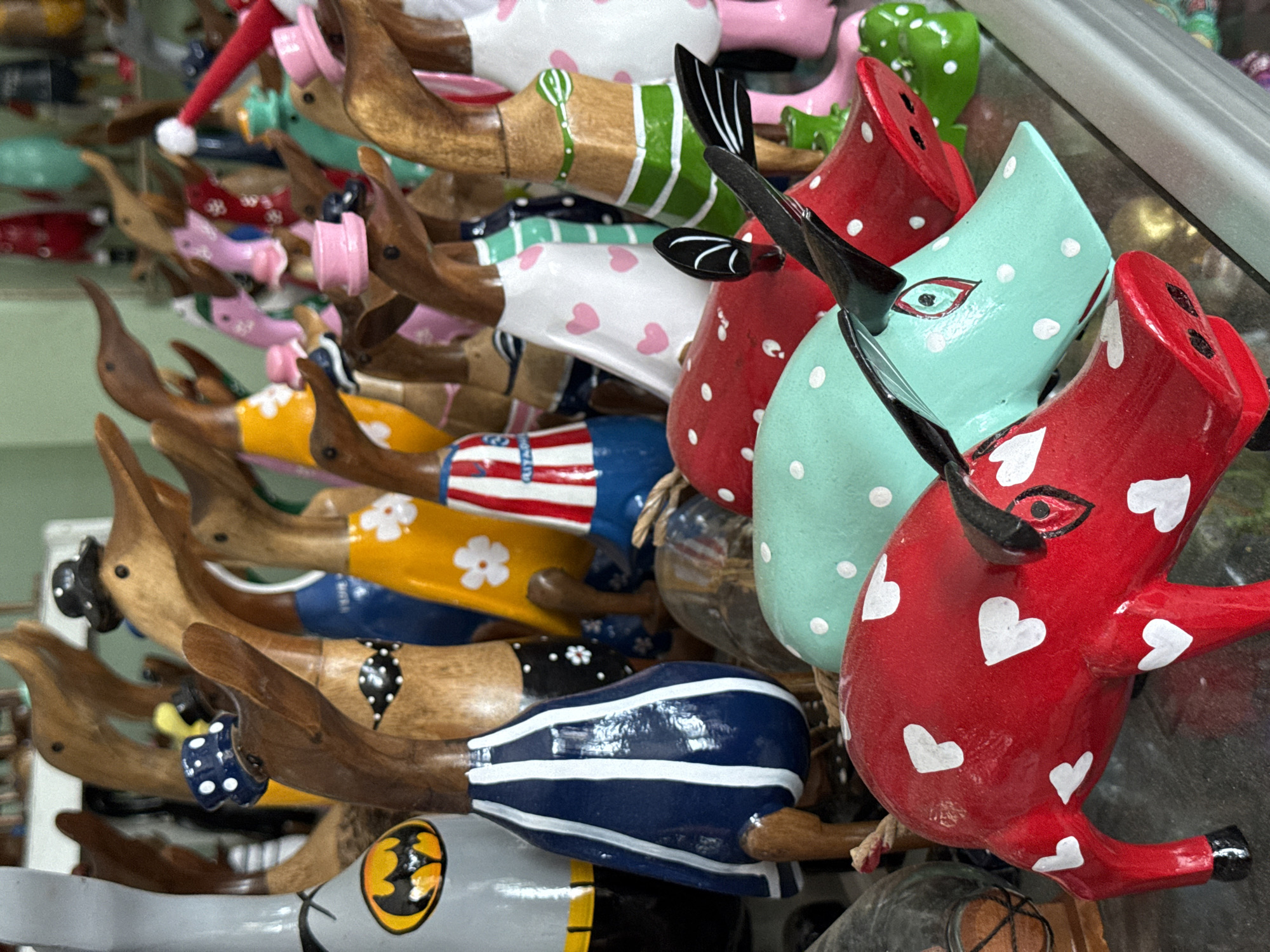 The image captures a dense display of numerous decorative wooden ducks or stylized bird figures, likely artisanal souvenirs. Each piece features a carved wooden head, beak, and legs, while the body is painted in vibrant colors with diverse patterns.
The ducks are brightly painted in a palette including red, white, blue, green, yellow, pink, and light blue/mint green, contrasted with the natural brown wood of their heads and limbs. Many bodies feature distinct patterns: white polka dots on red and mint green, pink hearts on white, white hearts on red, white stripes on blue, and green stripes on a natural wood background. Some ducks showcase specific themes, such as one with a miniature red Santa hat, another with a patriotic American flag design (red and white stripes), a yellow body with white floral patterns, and one sporting a yellow and black Batman logo.
The figures are closely packed and stacked, oriented in various directions, suggesting they are on display in a shop or market stall. The background is blurred, hinting at more items or the interior of a retail space, with subtle reflections possibly indicating a glass shelf or display case. No human activity is depicted, as the focus is entirely on the inanimate objects. There is no visible text, apart from the iconic Batman logo on one of the figures. The overall impression is one of a colorful, handcrafted collection offered for sale, consistent with the context of Ubud District, known for its arts and crafts.