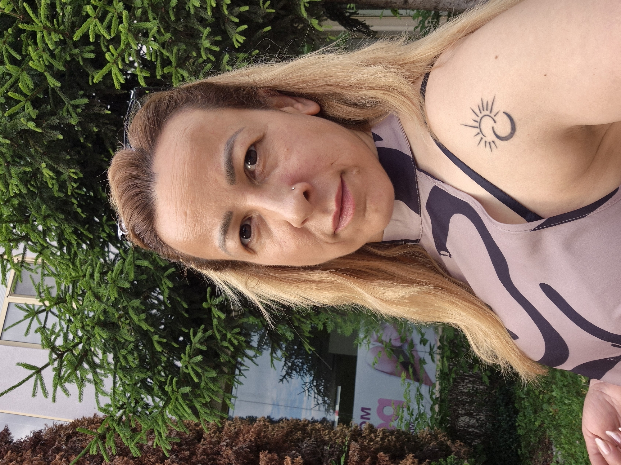 A medium-close-up outdoor portrait captures a woman with blonde hair, appearing to be in her 40s to 50s, looking directly at the camera. She has dark eyes, a small silver stud piercing on her left nostril, and light skin with visible facial lines and minor moles. Her long, blonde hair is styled back and falls over her shoulders. She wears a sleeveless, high-necked top featuring a light taupe background with a dark, abstract wavy pattern; a black bra strap is visible on her right shoulder. A black tattoo depicting a stylized sun with rays and a crescent moon, possibly interlocked, is visible on her upper right arm. A portion of her left hand with light-colored, manicured nails is visible in the lower-left corner.
The background consists of dense foliage and a building. To the far left, a tall, reddish-brown tree with textured bark stands. Behind the woman, a lush green conifer tree with distinct needles provides a natural backdrop. Further behind the trees, a modern building with large glass windows is visible. Reflections in the windows show parts of a blue sky with clouds and additional greenery. Through one of the windows, a partial sign with large pink/purple letters "OM" and "a" is discernible, along with a stylized graphic, suggesting a commercial establishment like a spa or salon. The scene is illuminated by natural daylight.