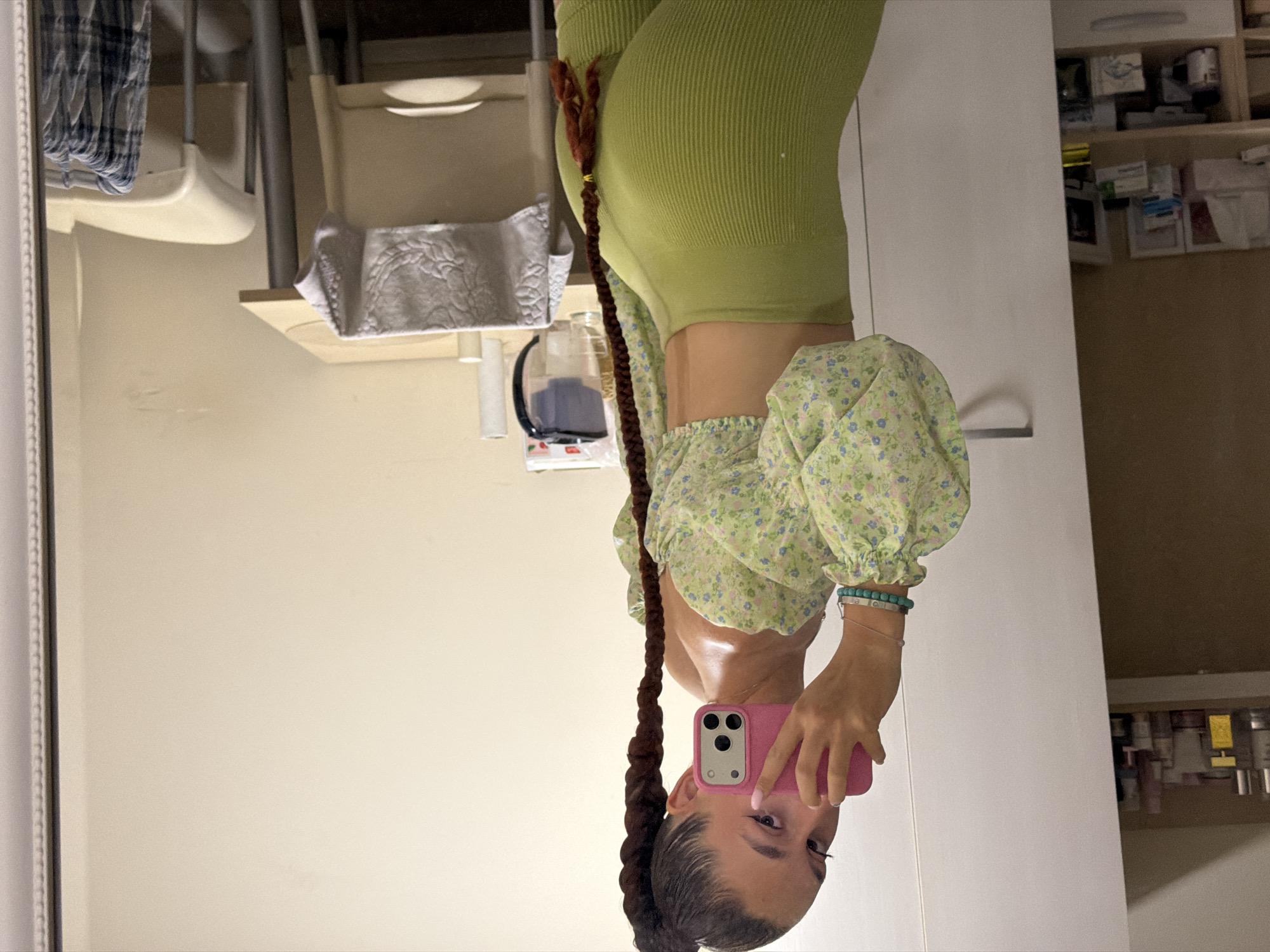 A young woman with dark hair in a long braid is taking a mirror selfie in what appears to be a bedroom or dressing area. She is wearing a light green off-the-shoulder top with a floral pattern and high-waisted green leggings. She is holding a pink smartphone to take the photo. In the background, to her left, is a white wardrobe with shelves containing various bottles and products. To her right is a small table with a chair, on which a folded white towel is placed. A white pitcher and a roll of paper towels are also visible on the table. The overall scene suggests a personal living space, possibly in Sofia, Bulgaria, given the request's context.