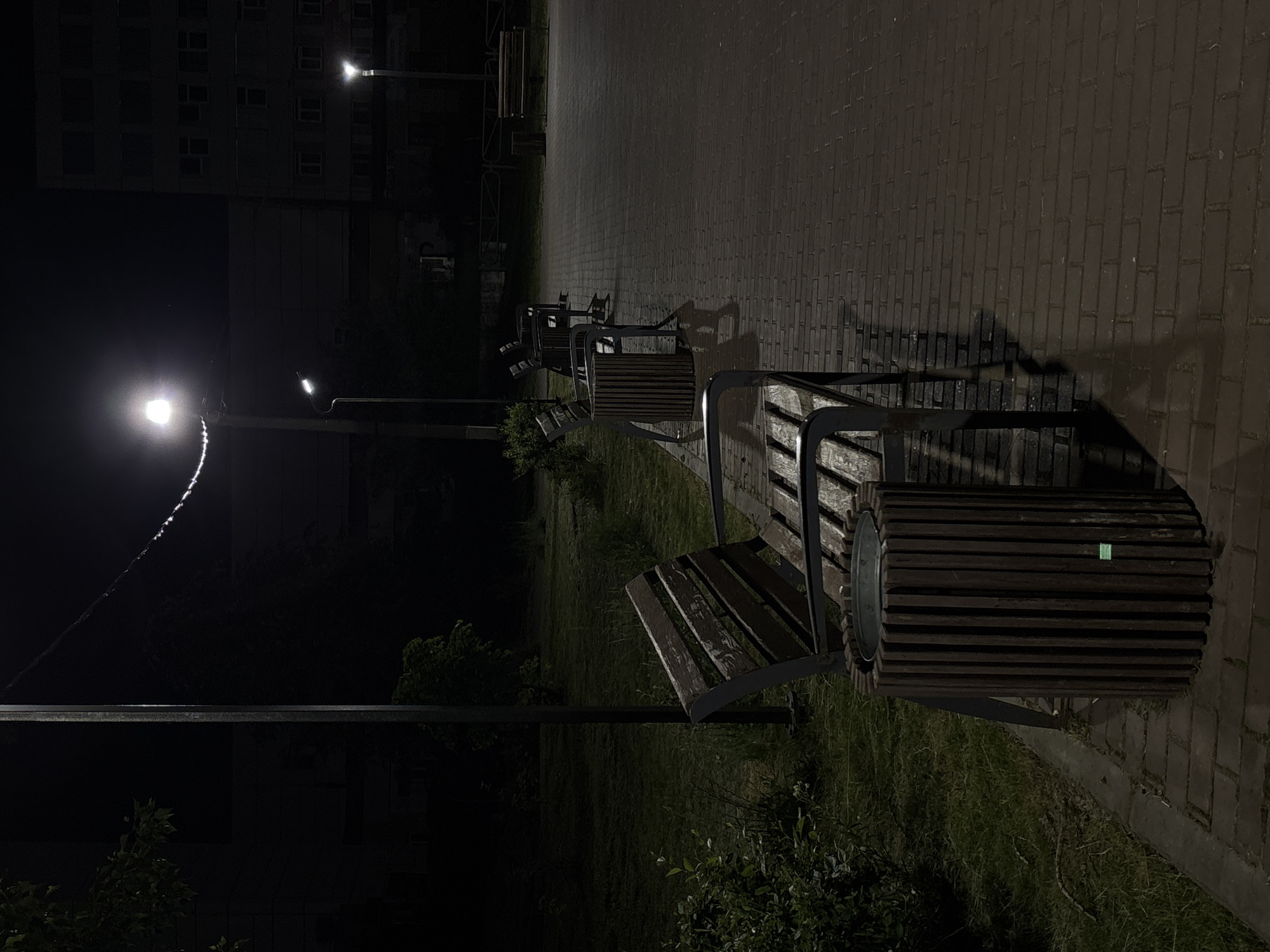 The image depicts a nighttime scene in Chaikovsky, Russia, likely in a public park or walkway. The primary focus is on a paved brick pathway illuminated by streetlights. Several park benches, constructed with wooden slats and dark metal frames, are positioned alongside the path, accompanied by cylindrical wooden trash receptacles. Green grass and sparse foliage are visible to the left of the pathway, bordering a dark, unlit area. In the background, a multi-story building with visible windows stands against the night sky, and other streetlights and power lines are present. The lighting creates long shadows across the path, indicating the time of night. No people are present in the image.