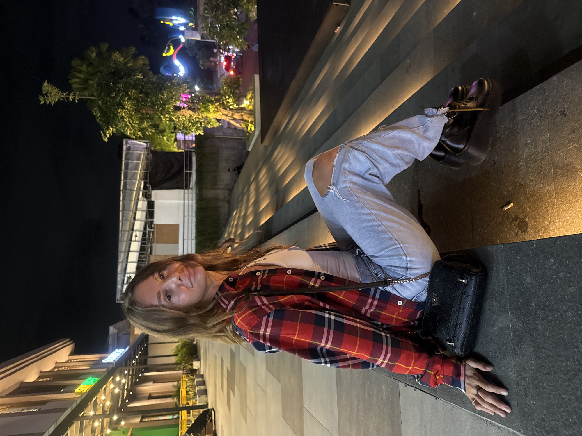 A woman is sitting on the edge of a series of steps. She is wearing ripped light blue jeans, a white top, and a red and blue plaid shirt. She is wearing black platform shoes. A black purse is next to her. The area appears to be an outdoor area in Kuta, Indonesia, at night. Lights are visible in the background.