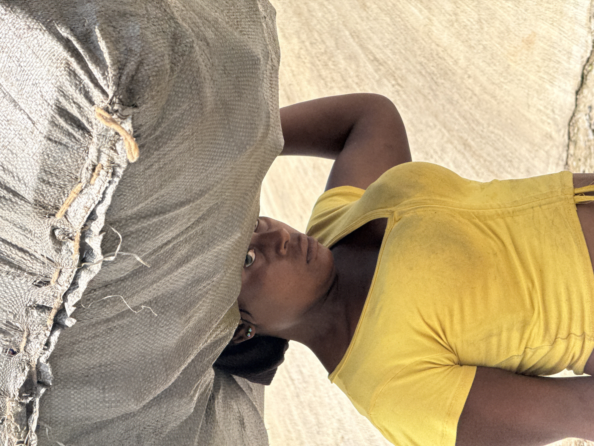 A woman wearing a yellow shirt is carrying a heavy sack on her shoulder. She is looking up at the camera. The location of the image is not available.