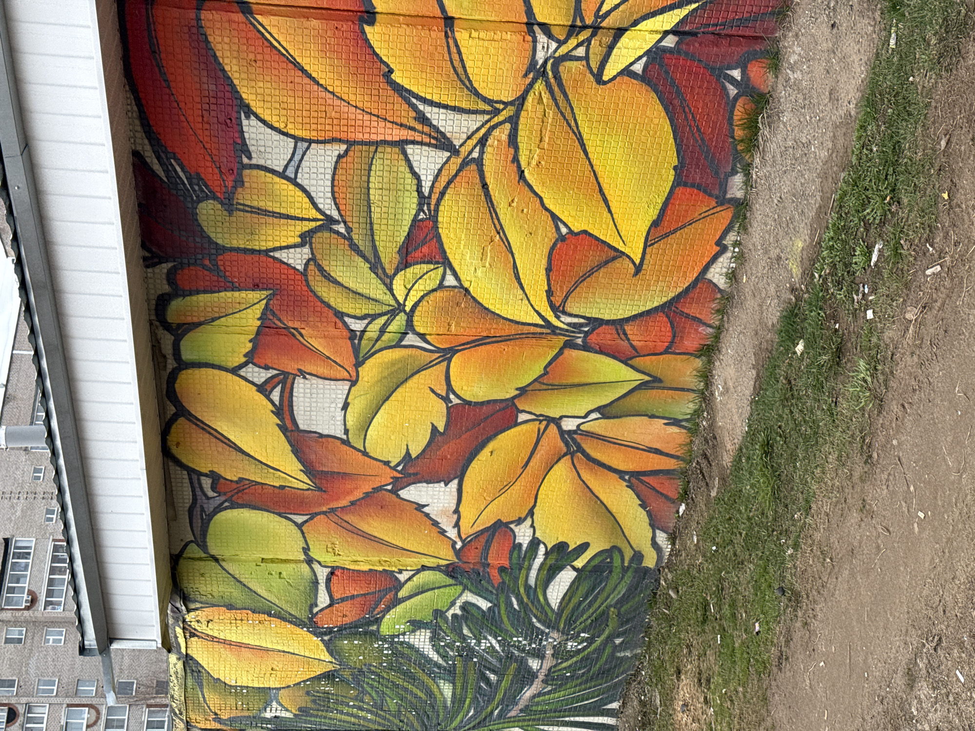 The image depicts a mural of autumn leaves painted on a wall. The leaves are primarily yellow, orange, and red, with black outlines. The mural appears to be on a tiled wall, and a white building is adjacent to the mural on the left side of the image. To the right of the mural, there is a patch of grass and a dirt path. The location of the mural is in the city of Unknown, Unknown.