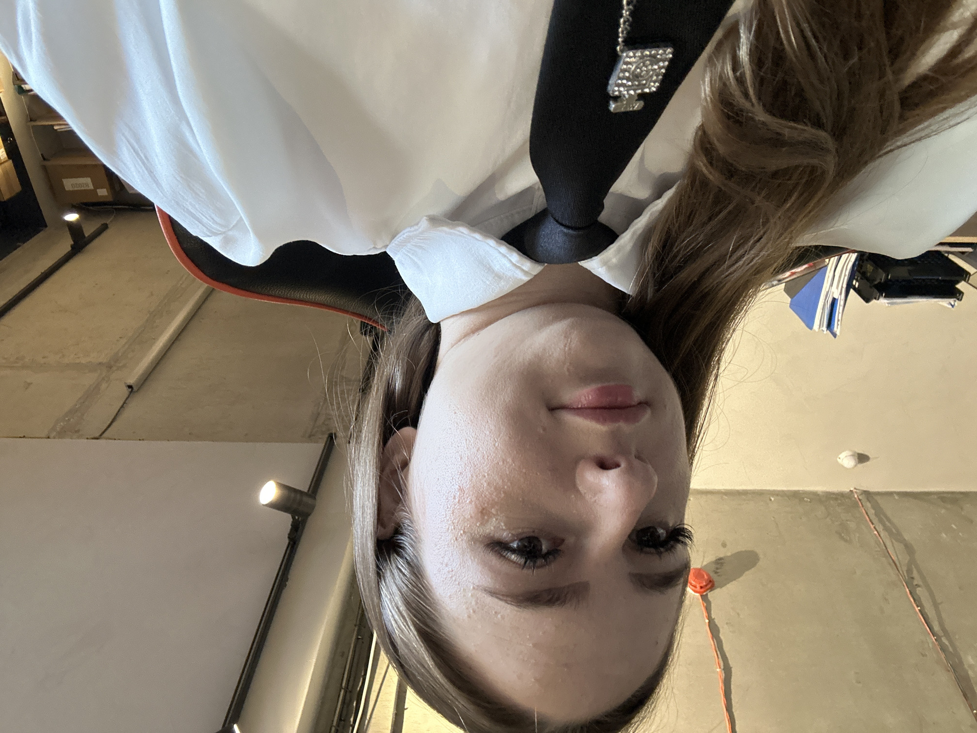 A medium shot, taken from a low angle, shows a young woman with long, brown hair and fair skin. She is wearing a white collared shirt and a black tie, which is adorned with a silver-toned, rectangular charm resembling a perfume bottle, encrusted with small, clear stones. The woman's head is tilted slightly upwards and to her left, and her gaze is directed towards the camera. Her makeup includes dark eyeliner and mascara, and her lips are painted with a neutral shade.
The background is an indoor environment. To the left, a black office chair is visible, with an orange trim. Behind the chair, stacks of folders and papers are arranged on a desk. The ceiling is exposed concrete, with visible wiring and industrial lighting fixtures, one of which is illuminated. The overall lighting is bright, casting some shadows on the concrete ceiling. The setting appears to be an office or a workspace.