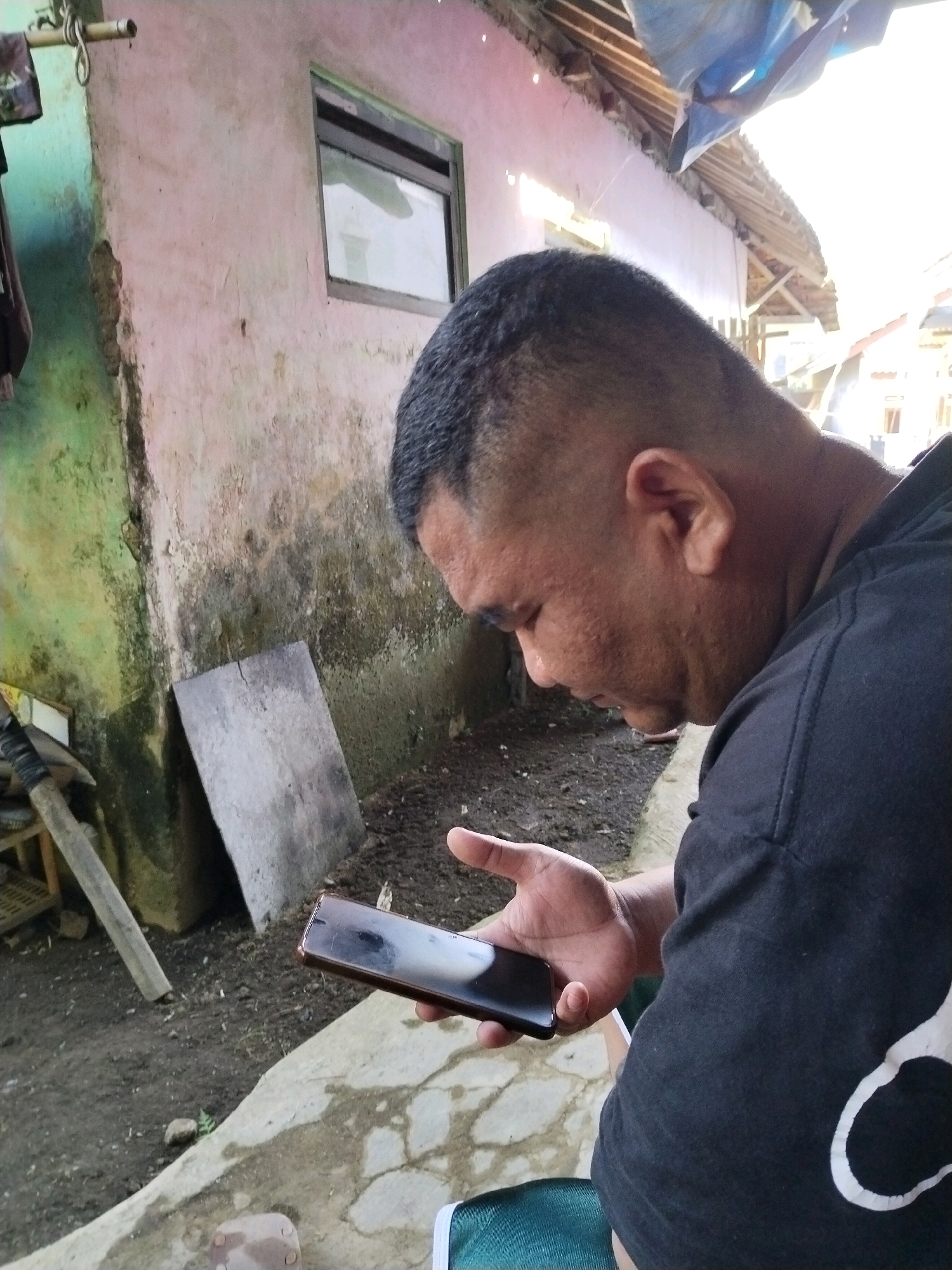A close-up, outdoor, eye-level medium shot captures a man in Desa Cidugalen, Indonesia, looking down at a dark-colored smartphone held in his right hand. The man, seen from the chest up and in profile, has a short buzz cut and is wearing a black t-shirt with a white graphic design on the left side of the chest. His skin appears oily, and he has visible pores on his face. He is positioned in front of a pale pink wall with a green section on the left, showing signs of age and wear, including cracks and patches of discoloration. A rectangular window with a dark frame is visible on the pink wall. To the man's left, a flat, gray object, possibly a stone slab, leans against the wall. The ground is composed of dirt and concrete. Overhead, part of a corrugated metal roof with a blue tarp draped over it is visible. The scene is illuminated by bright sunlight, creating a high-contrast environment.