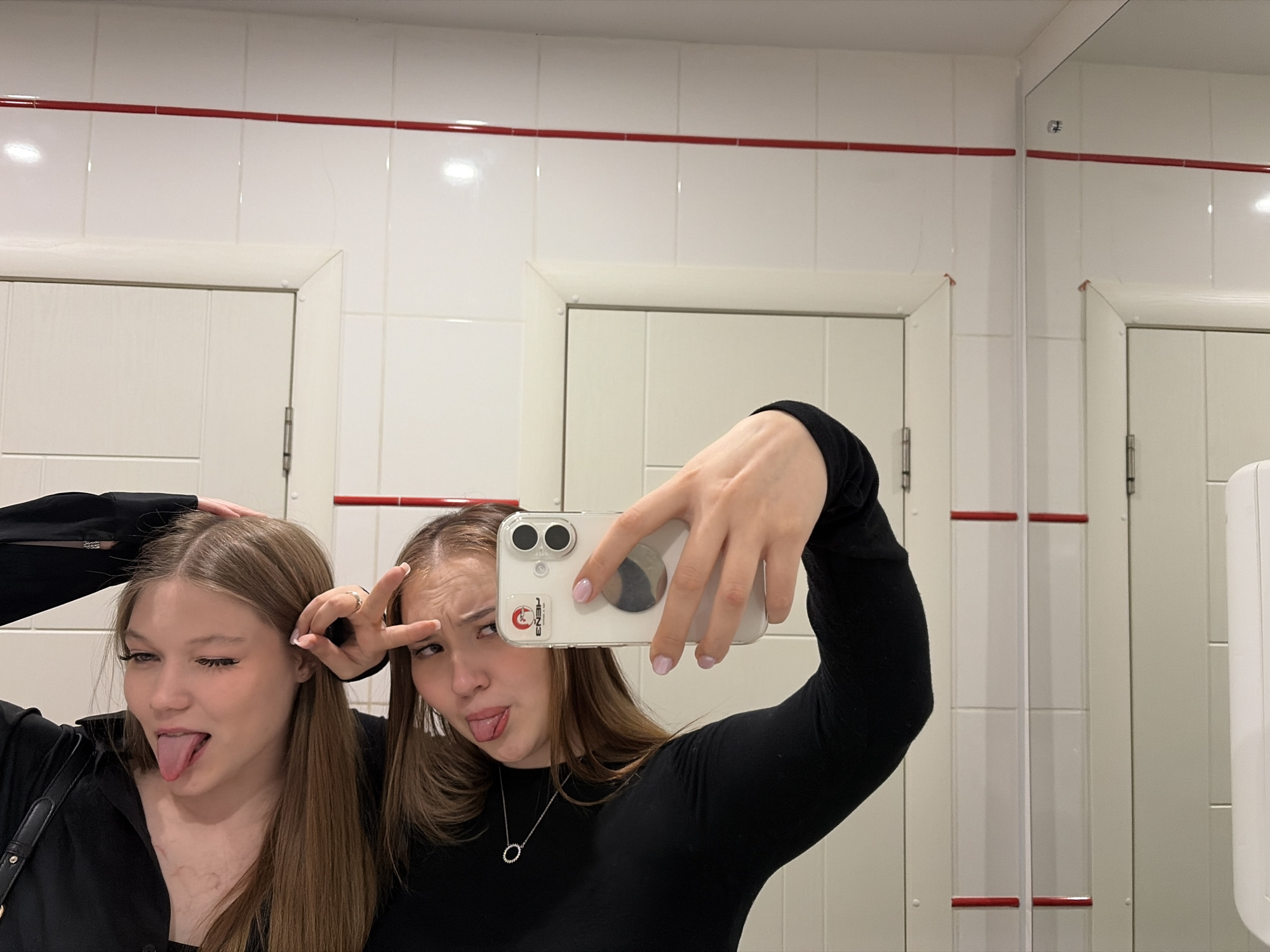Two young women are taking a selfie in what appears to be a public restroom. The restroom has white tiled walls with a red stripe near the top. There are at least two doors and a mirror visible. The women are making silly faces. One woman is sticking out her tongue, while the other is sticking out her tongue and holding up a peace sign near her eye. The location is Chaikovsky, Russia.