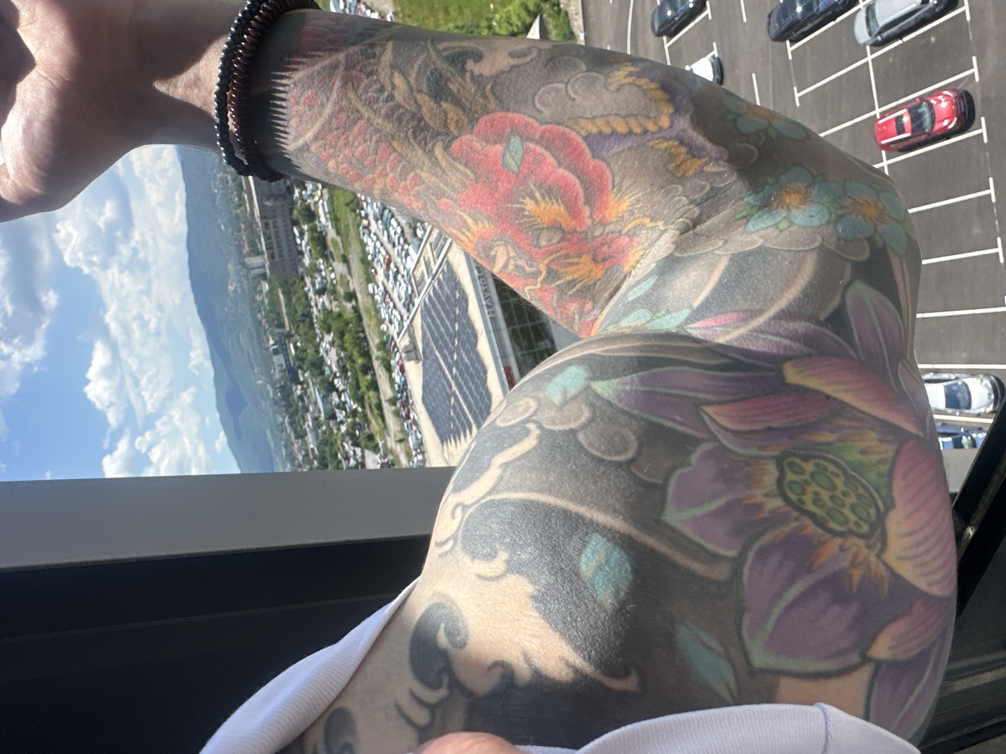 The image shows a close-up of a person's tattooed arms against a backdrop of a city, specifically Sofia, Bulgaria. The tattoos feature large, colorful designs including lotus flowers and a dragon. The person is wearing a white shirt. The background reveals a panoramic view of a city with buildings, roads, and a large parking lot filled with cars. Mountains are visible in the distance under a cloudy sky. A partial view of a building with the text "HAVTO" is discernible. The perspective suggests the photo was taken from a high vantage point, likely a building overlooking the city.