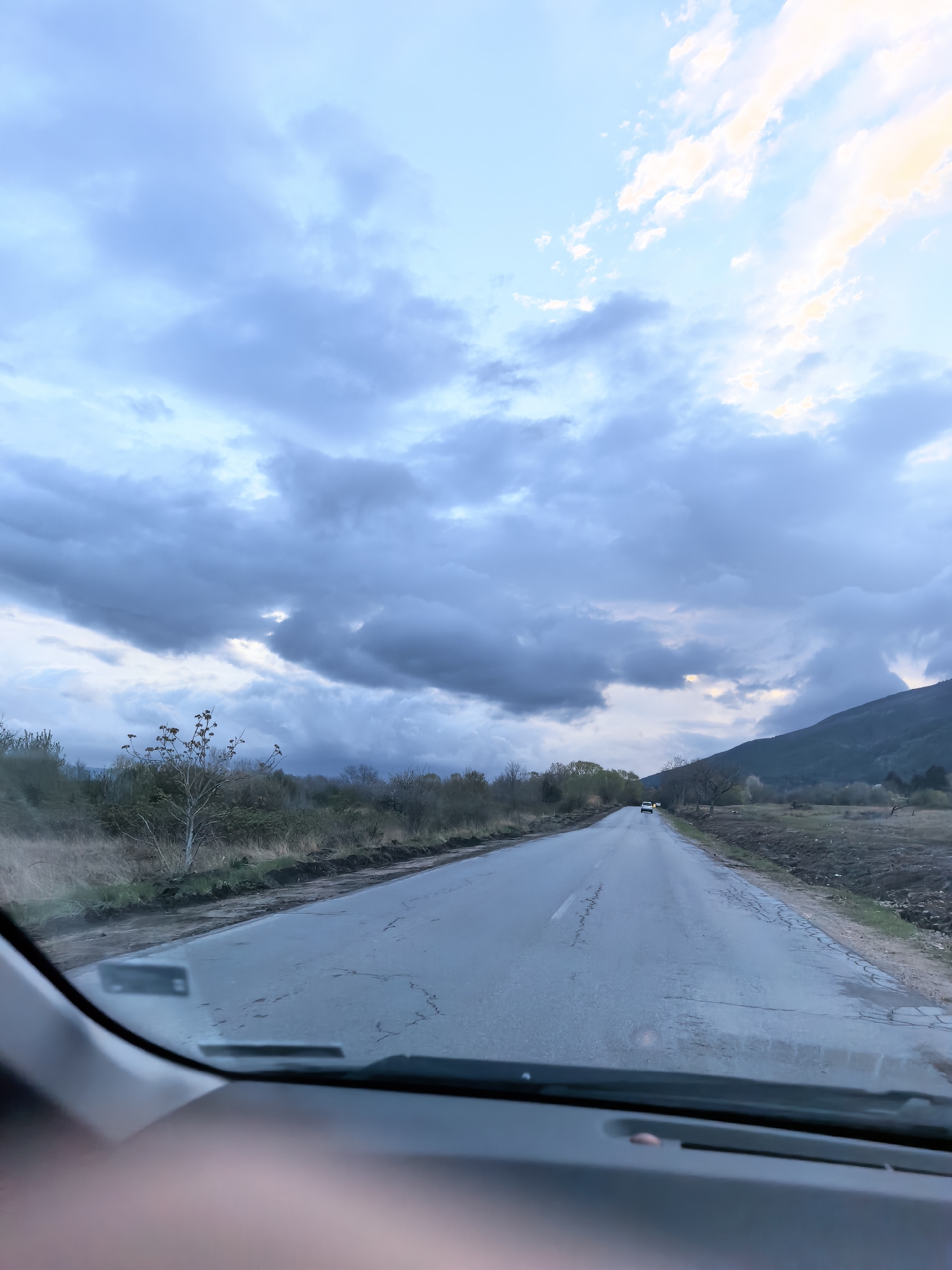 The image presents a view from inside a vehicle, looking out through the windshield onto a two-lane road that extends into the distance. The road surface appears wet or damp, with visible cracks in the asphalt, suggesting recent rain or high humidity. On both sides of the road, there's a rural landscape featuring sparse vegetation, including dry grasses, bare bushes, and a few small trees.
The sky is a prominent feature, filled with dramatic, heavy grey and dark blue clouds that stretch across the frame. However, patches of lighter blue sky are visible, and certain clouds, particularly towards the upper right and center, are illuminated with a soft, warm yellowish-white light. This interplay of dark clouds and brighter light suggests either sunrise or sunset, with the sun attempting to break through the overcast conditions. In the far distance on the right, a dark, undulating mountain or hill range rises against the horizon.
Two other cars are visible further down the road: a white vehicle is clearly seen, followed by another car whose yellowish headlights are discernible. The dashboard of the viewing vehicle occupies the lower part of the frame, with a blurry, indistinct shape, possibly a hand or arm, in the lower-left corner. The overall impression is of a tranquil journey through a natural landscape under a striking and somewhat dramatic sky. No identifiable text is visible within the image.