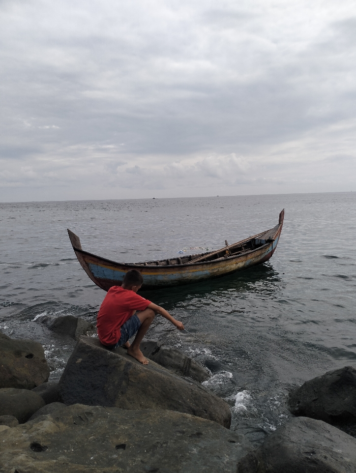 A boy in a red shirt and blue shorts sits on gray rocks near a body of water. His right arm extends toward the water, and he appears to be holding a fishing line. Behind him, a small wooden boat floats in the water. The sky is overcast with gray clouds. The water extends to the horizon, and some distant objects are visible on the water. The foreground consists of large, dark rocks.