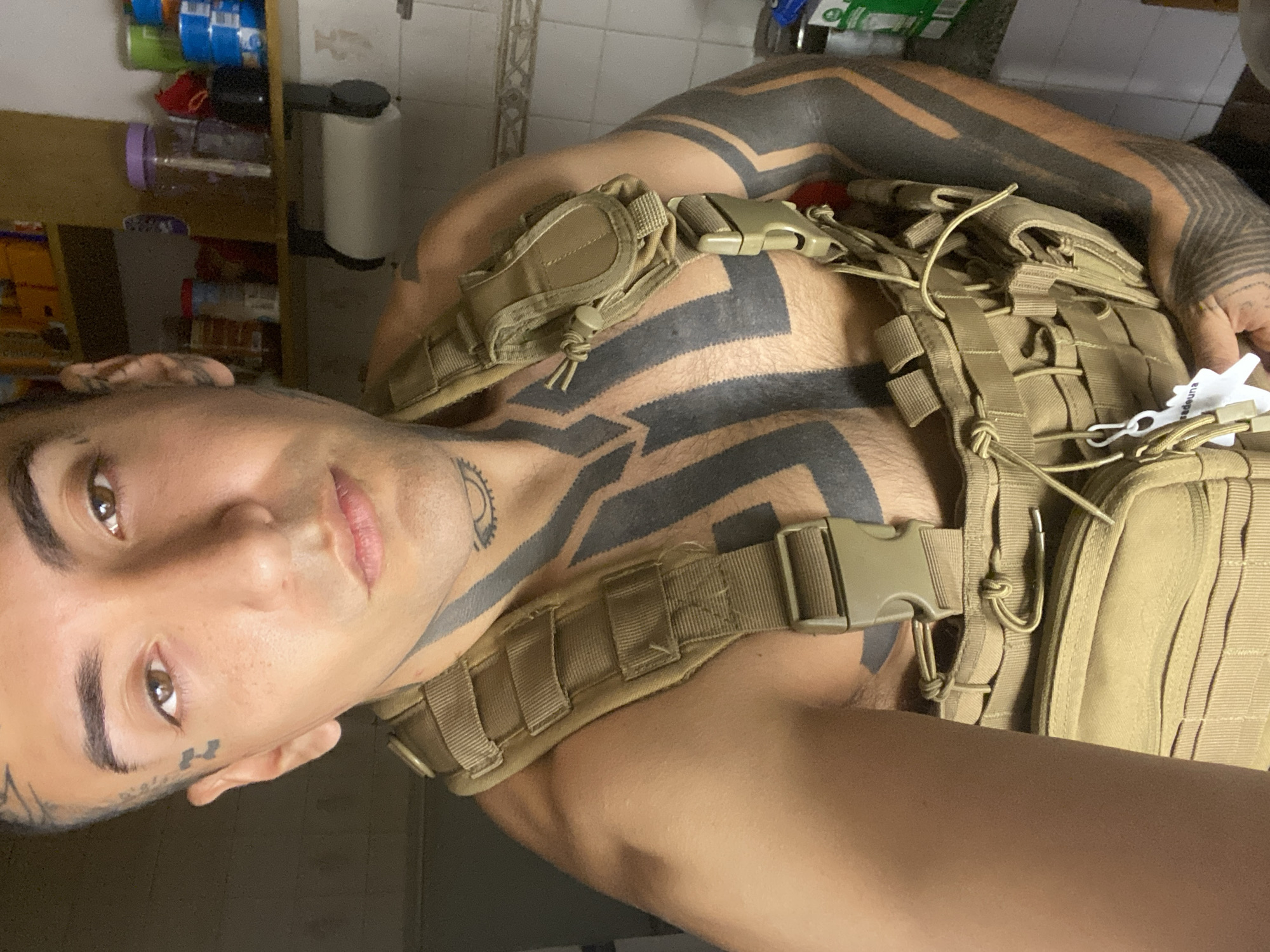 A fair-skinned individual with dark hair is wearing a tactical vest. The person has prominent black tattoos on their arms, chest, and neck. Additional tattoos are visible on the face near the temple and below the right eye. The vest is beige, with multiple straps and buckles. The background shows a portion of a shelf with various items, including food containers and a white dispenser. Without additional information, determining the exact location in Buenos Aires, Argentina, is impossible.