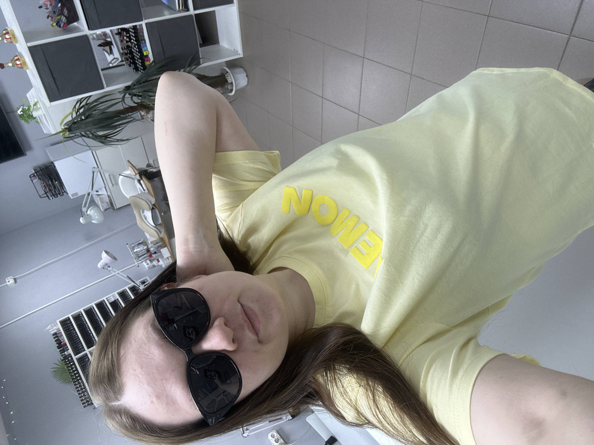 A young woman wearing a yellow t-shirt with the word "LEMON" printed on it and black sunglasses is taking a selfie. She is indoors, possibly in a salon or similar business, given the shelving unit visible in the background, which contains various items including what appear to be nail polish bottles. A plant sits on top of the shelving. The setting appears to be a modern, well-lit interior with light gray walls and tile flooring. The location is suggested to be Chaikovsky, Russia, though this cannot be confirmed from the image alone.