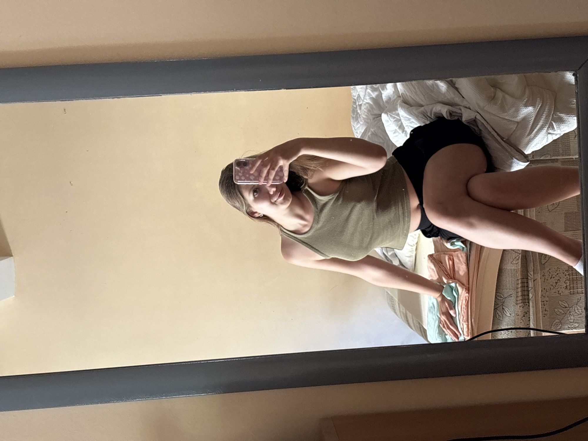 A young woman with light brown hair takes a mirror selfie while sitting on a bed in what appears to be a bedroom. She is wearing a green tank top and black shorts. Her left hand is holding a phone with a pink patterned case, and her right hand is resting on the bed. The bed is made with white bedding. To the left of the bed, there are folded pastel-colored cloths. The wall behind the bed is a light beige color. The room is framed by a large mirror with a dark gray frame. The image does not provide enough information to determine the specific location within the city of Bečići, Montenegro.