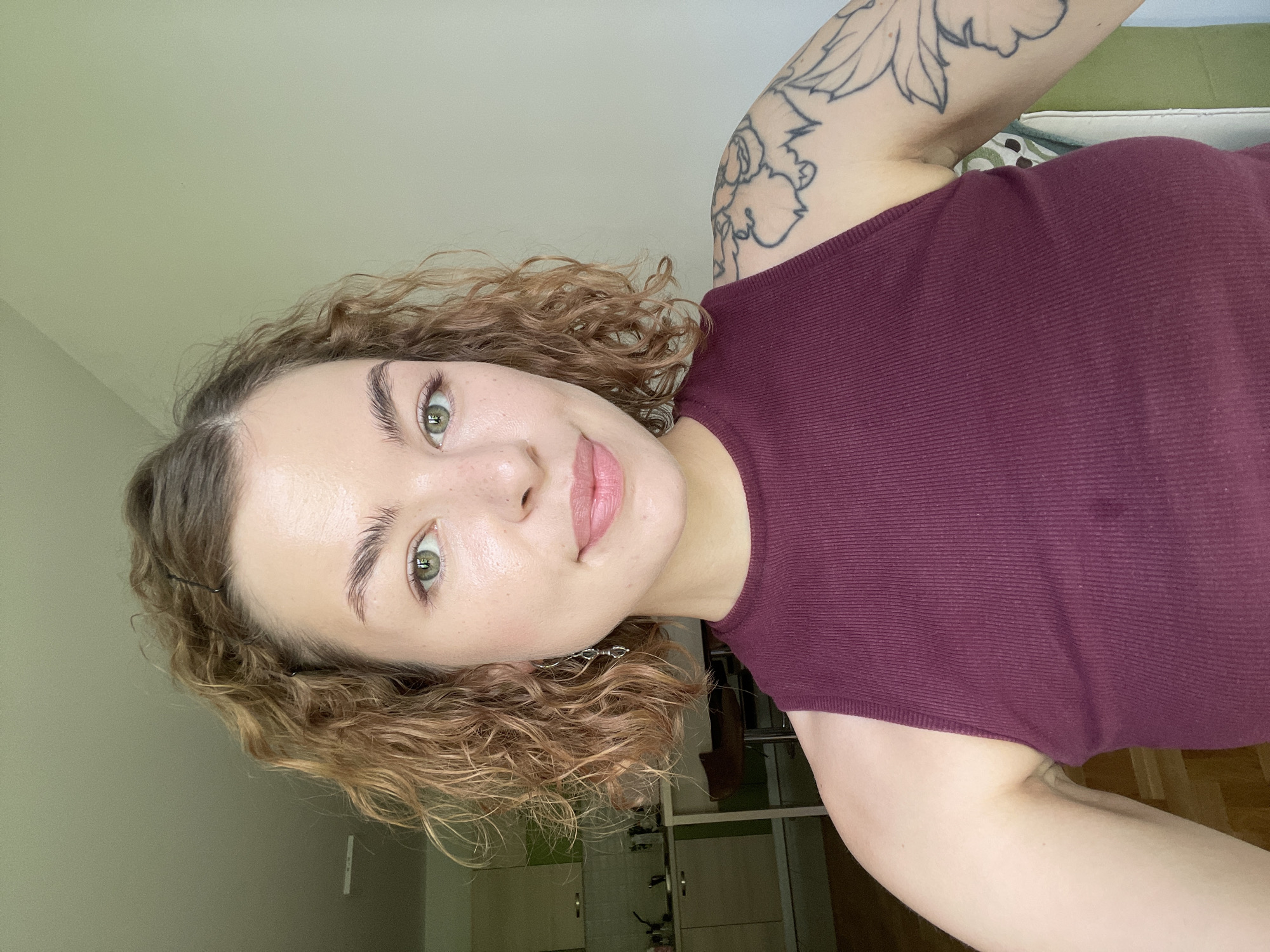 A woman with curly brown hair and green eyes is facing the camera. She is wearing a burgundy, sleeveless top. Her left arm has a floral tattoo. The background includes a light green wall, a cabinet, and a wooden floor. The location information is not depicted.