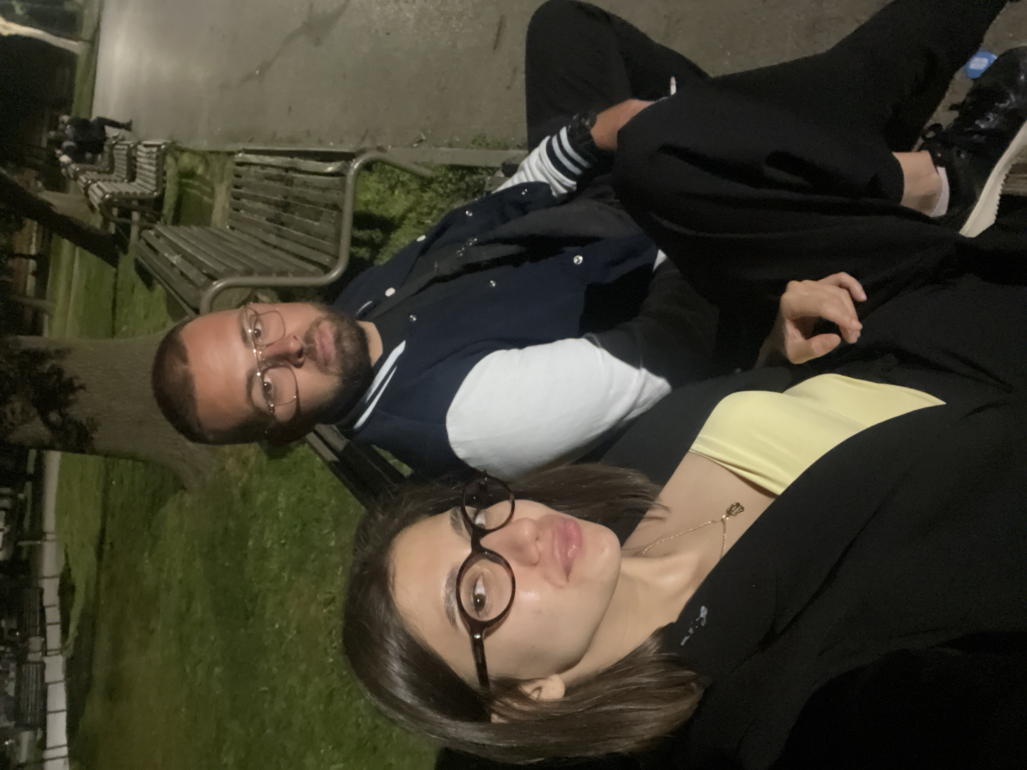 An outdoor, low-light shot captures two individuals relaxing in what appears to be a park in Sofia, Bulgaria, during the evening or night. The image is oriented sideways, with subjects appearing to lie down or recline, looking towards the camera.
The foreground features a woman with shoulder-length dark hair, wearing dark-framed round glasses, a light yellow strapless top, and a dark blazer or jacket. A delicate gold-colored necklace with a small pendant adorns her neck. Her expression is calm, with full lips.
Beside or slightly behind her is a man with a short beard and mustache, and short dark hair. He wears clear-framed glasses and a navy blue bomber-style jacket with white sleeves and striped cuffs, over a white inner layer, paired with dark pants and black sneakers with white soles. He also appears relaxed, looking towards the camera. His left arm is bent, revealing a watch on his wrist.
The background shows dark green grass, a concrete path, and several empty metal park benches, indicating an outdoor, public setting. The overall lighting is dim, suggesting late evening or nighttime conditions, with no strong artificial light sources immediately visible. No visible text is present in the image.