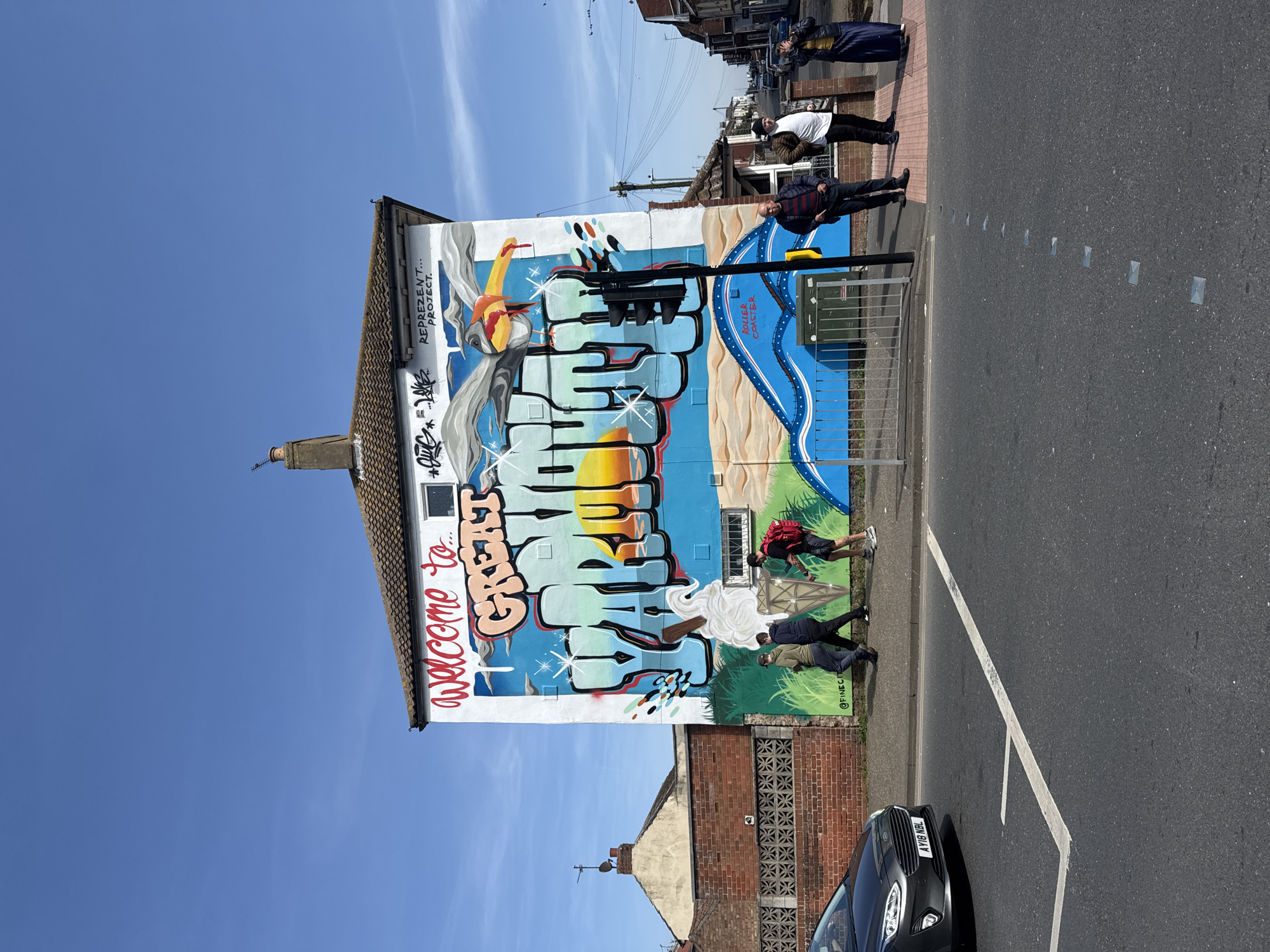 The image shows a street scene in Great Yarmouth, United Kingdom. A tall building is prominently featured, its side entirely covered in a large, colorful mural. The mural includes the words "Welcome to Great Yarmouth" in stylized graffiti lettering, accompanied by other vibrant imagery and artistic designs. Several people are walking on the sidewalk adjacent to the mural. A street with painted parking lines runs in front of the building, and a dark-colored car is parked at the curb. Other buildings are visible in the background, across the street. The sky is blue with scattered clouds.